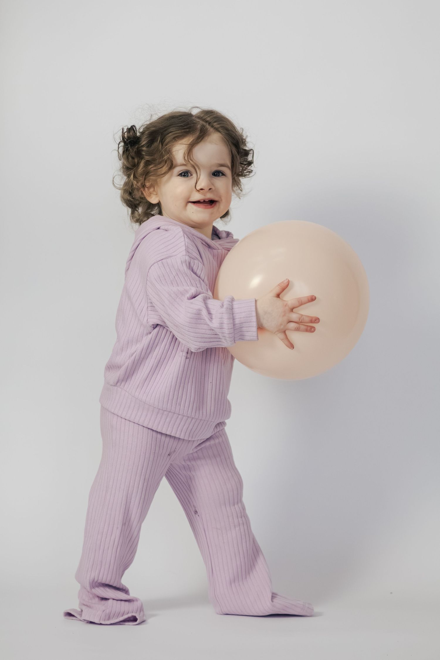 Adorable toddler girl in pink ribbed pajama set playing with a peach balloon on white background.