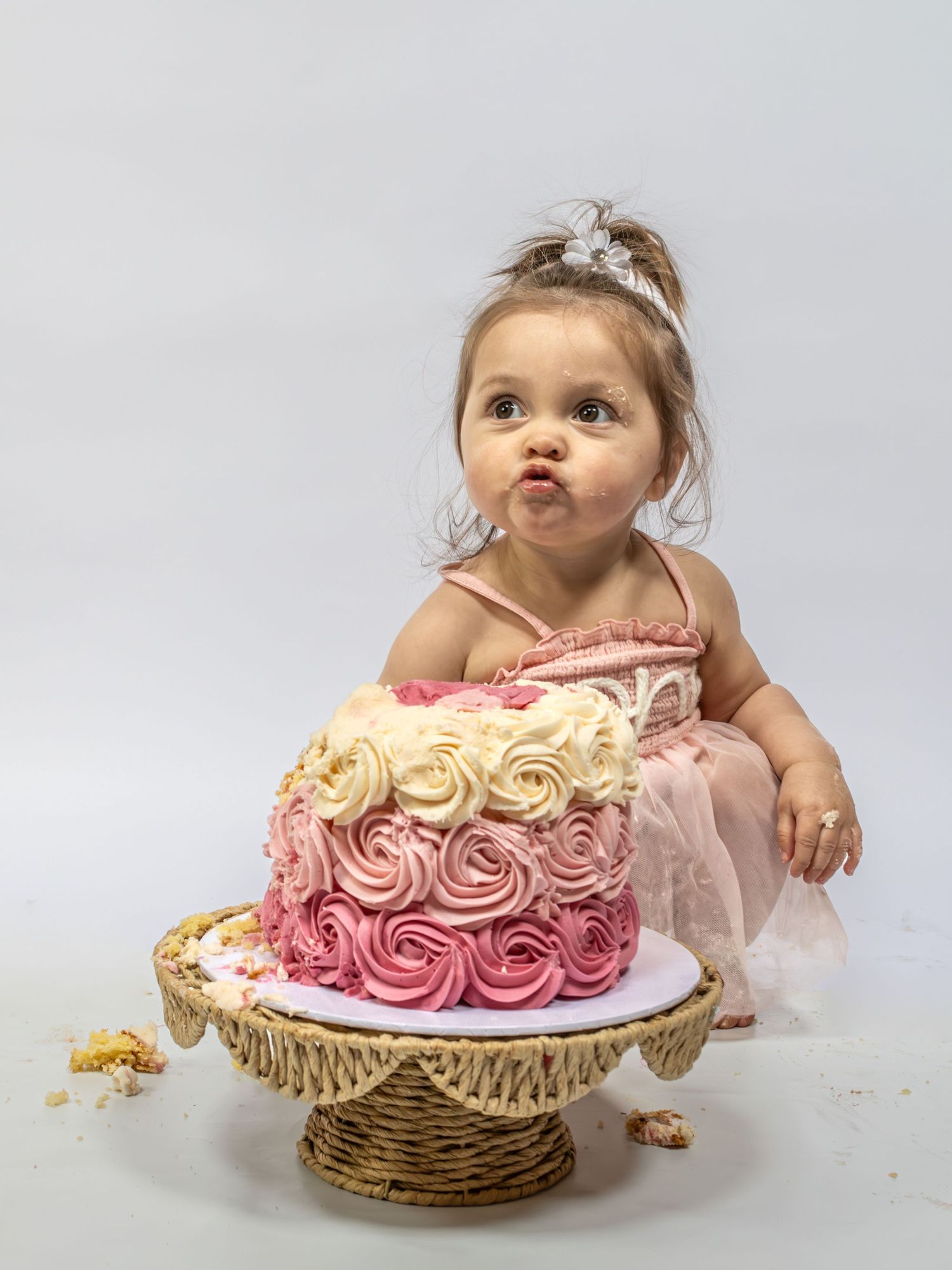 Cute baby girl in pink tutu and floral tiara posing with a pink ombre rosette smash cake on a wicker stand.