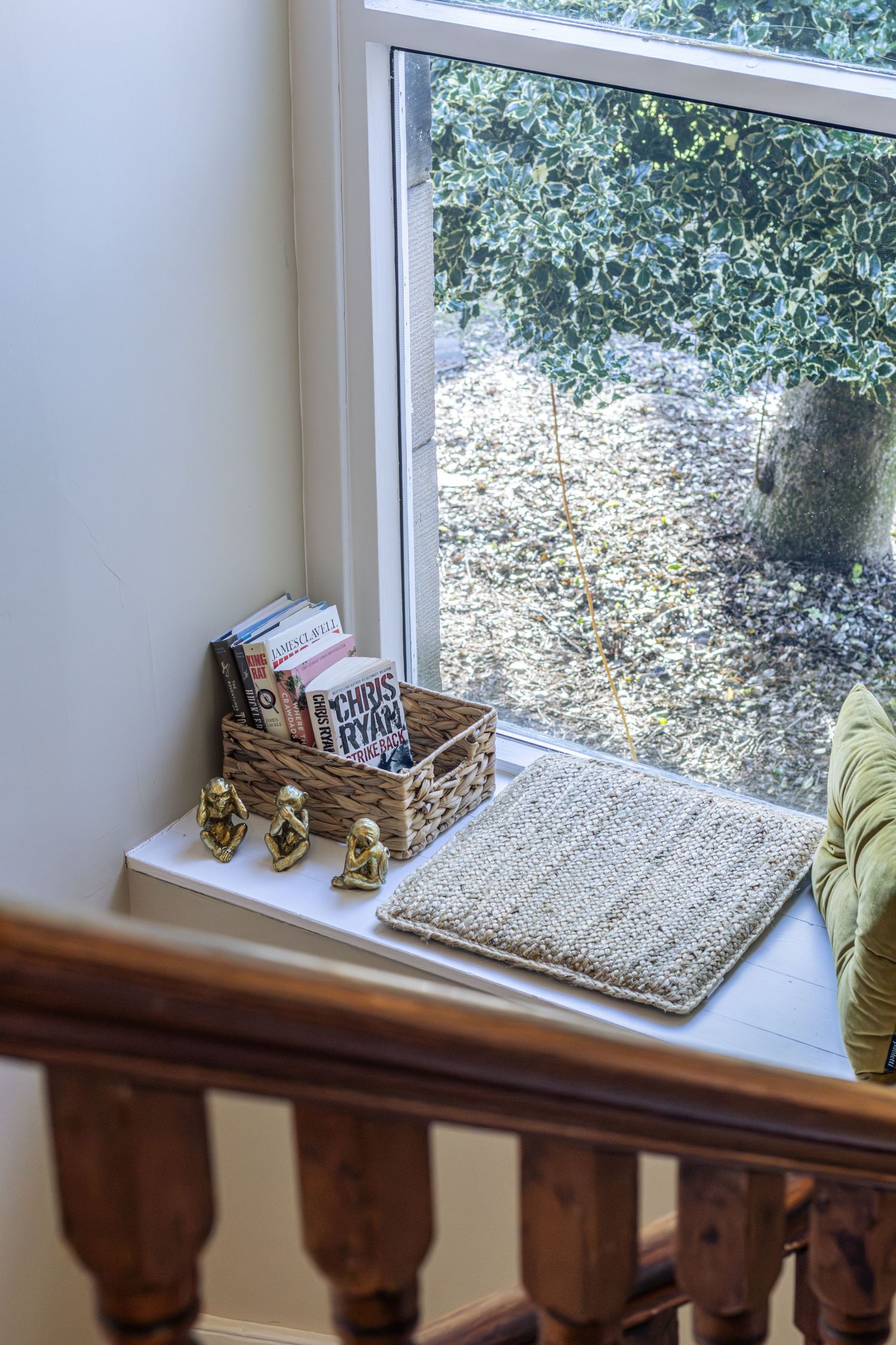 Cozy window seat nook with wicker basket of books, knitted cushion, and gold figurines overlooking a garden.
