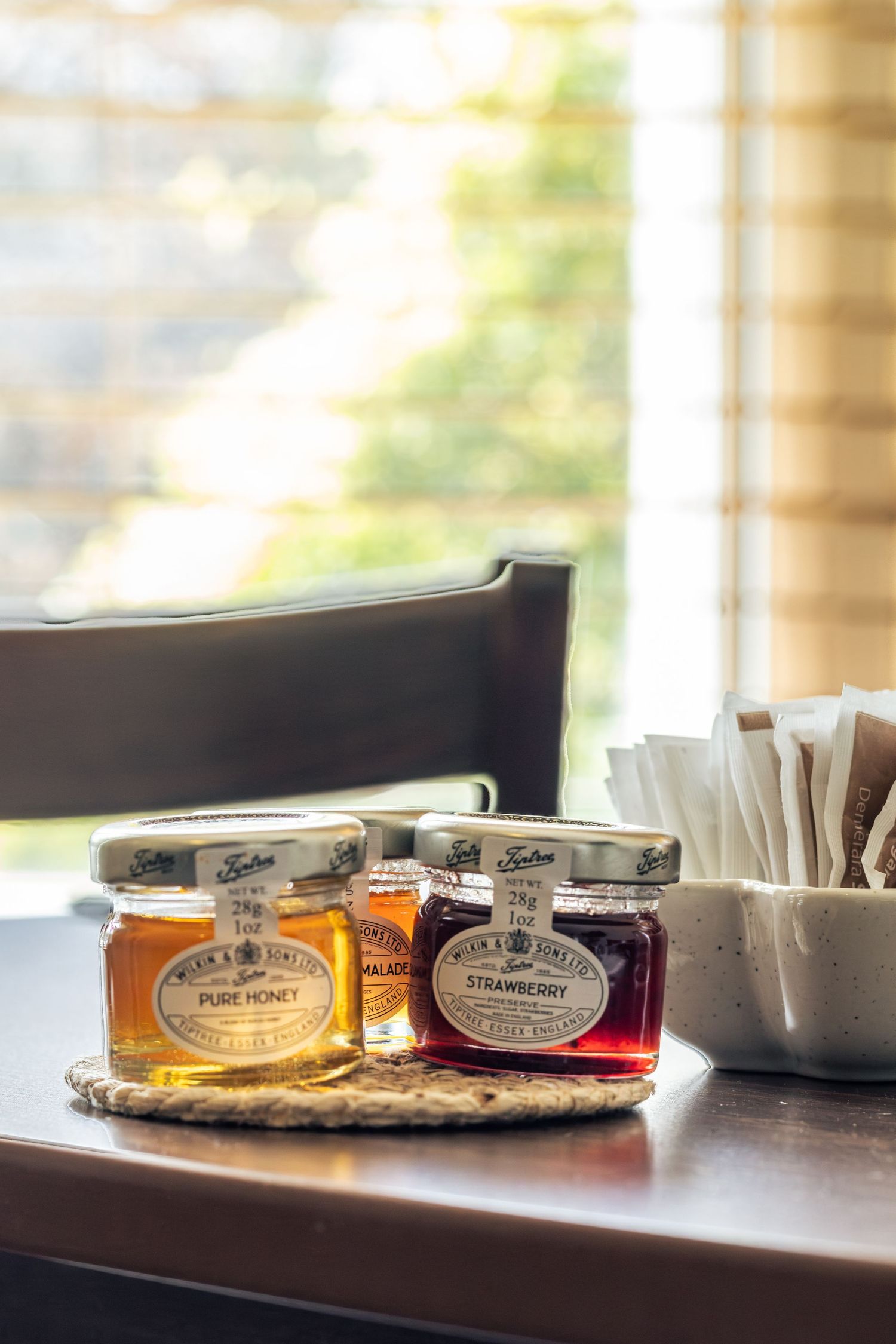 Three Tiptree mini jam jars including pure honey, marmalade, and strawberry on a wicker mat beside sugar packets.
