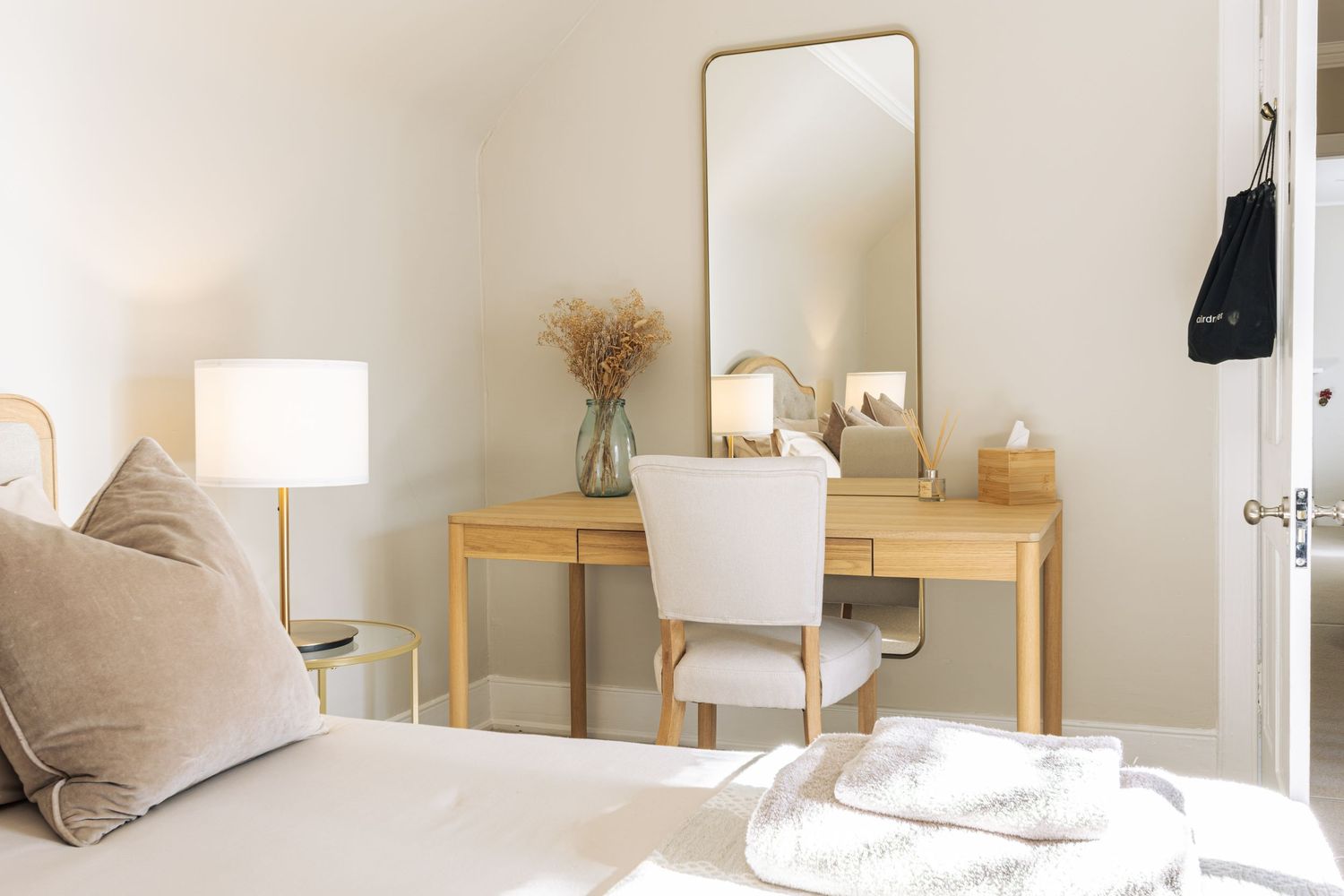 Minimalist bedroom vanity area with wooden desk, white chair, gold mirror, and lamp in a bright, neutral-toned room.