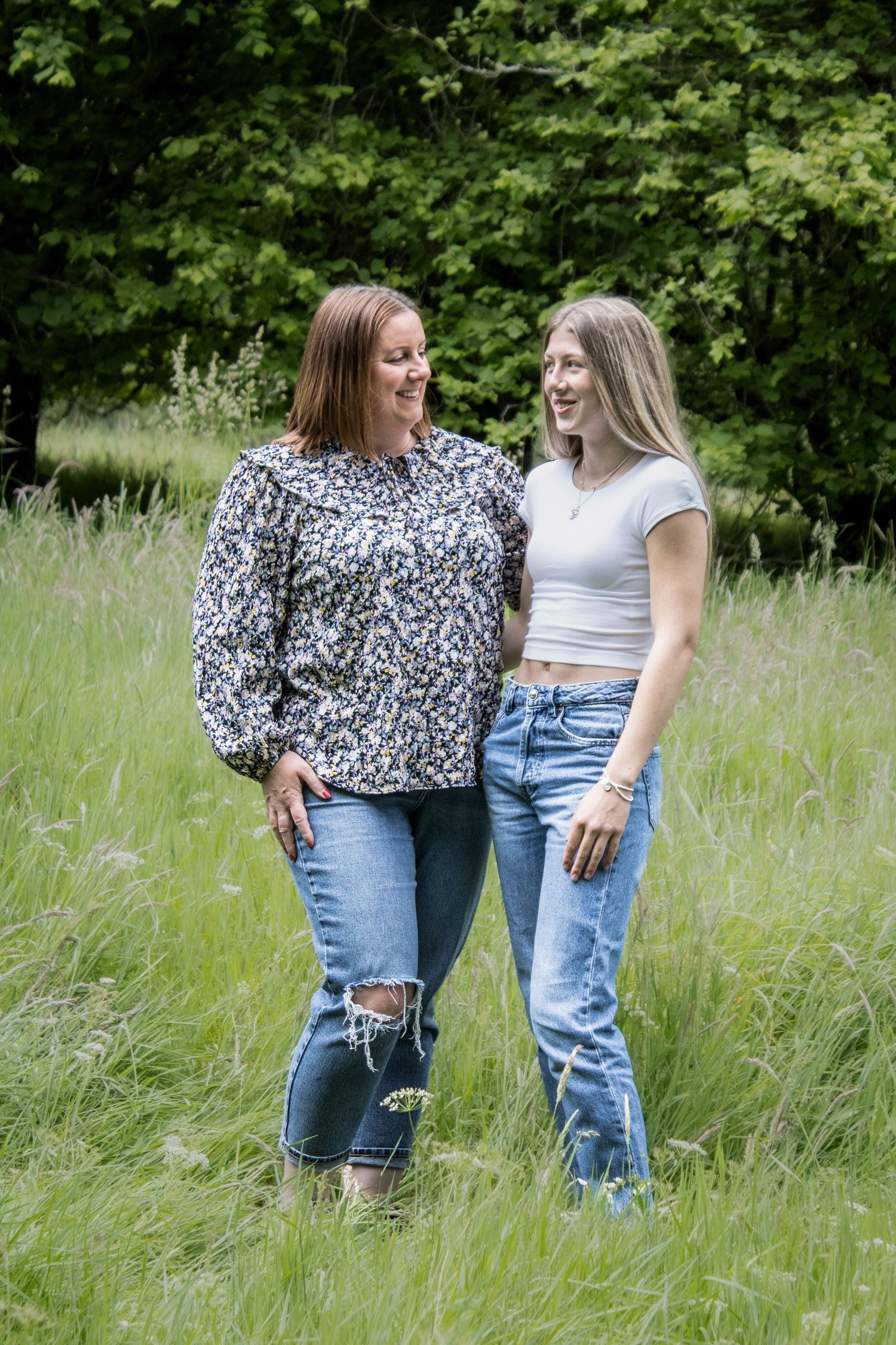 Two friends stand together in a sunny meadow wearing casual denim and summer attire.