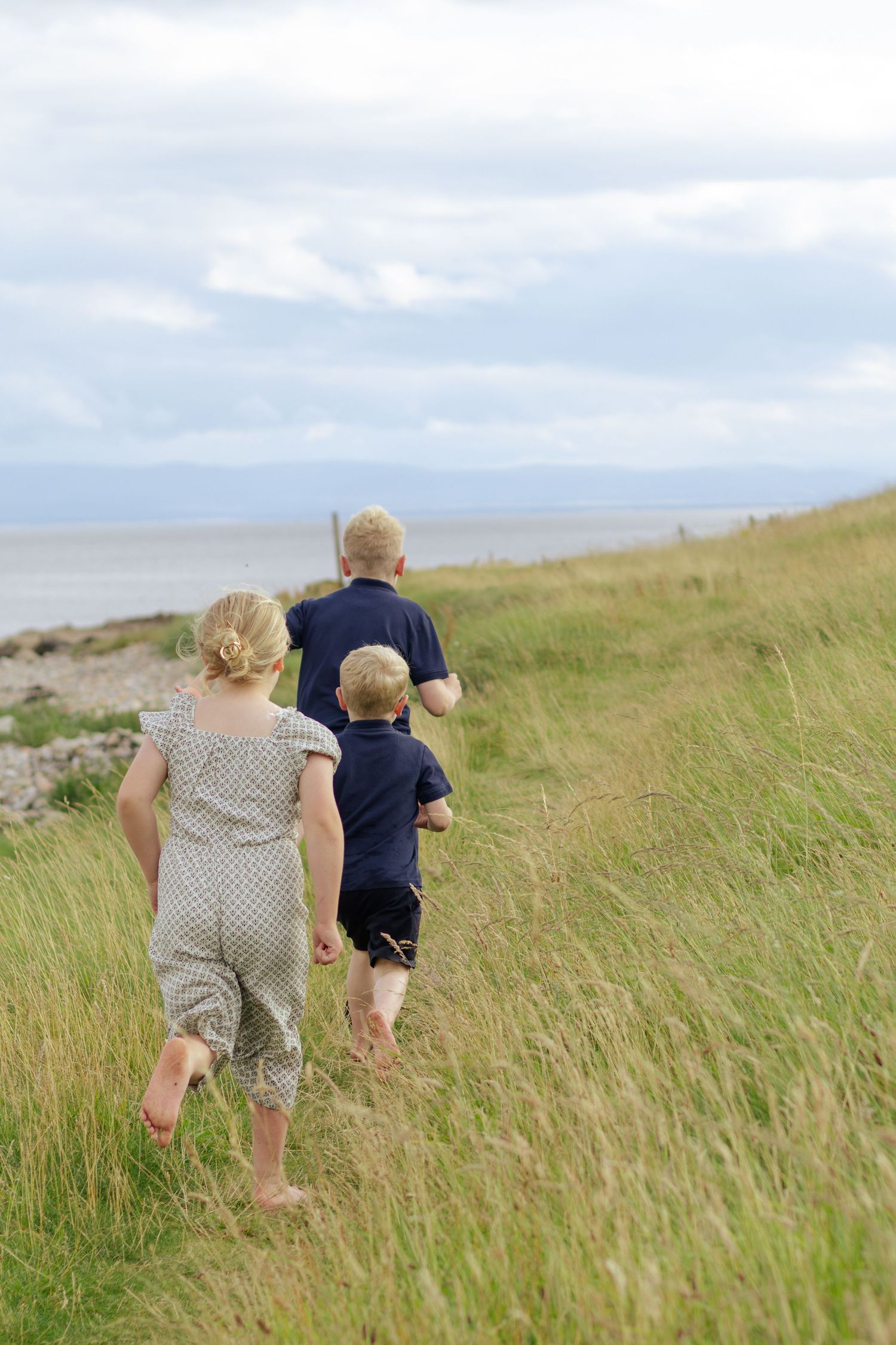 Three barefoot children running through tall grass toward a scenic coastal shoreline under a cloudy sky.