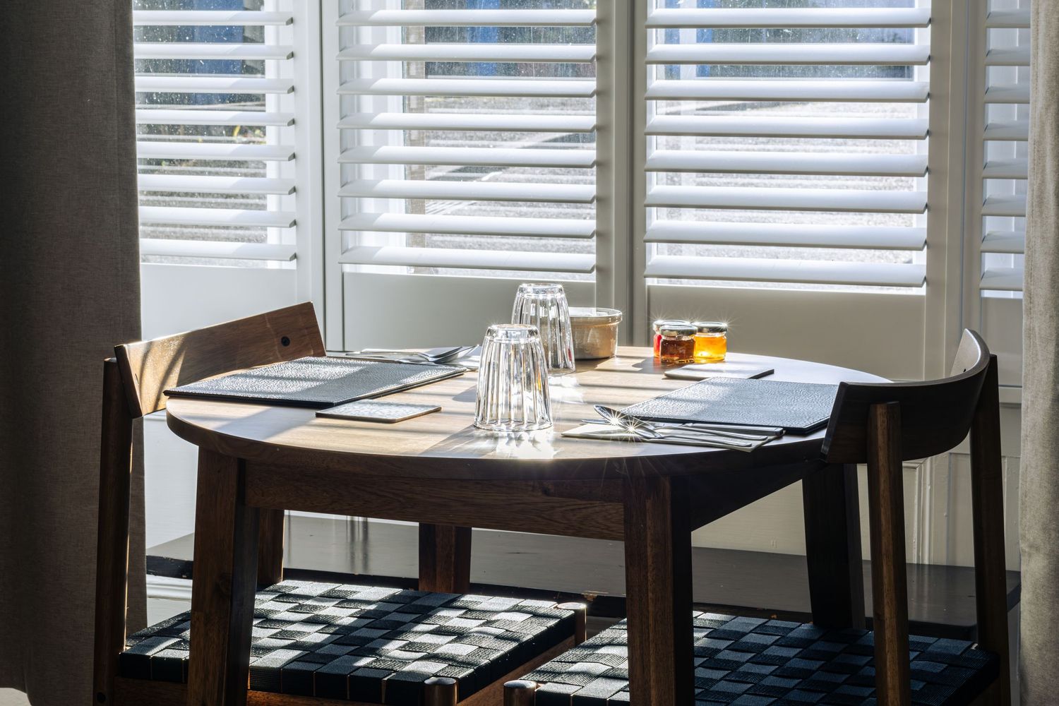 Sunlit restaurant table set with menus, glassware, and drinks beside white plantation shutters.