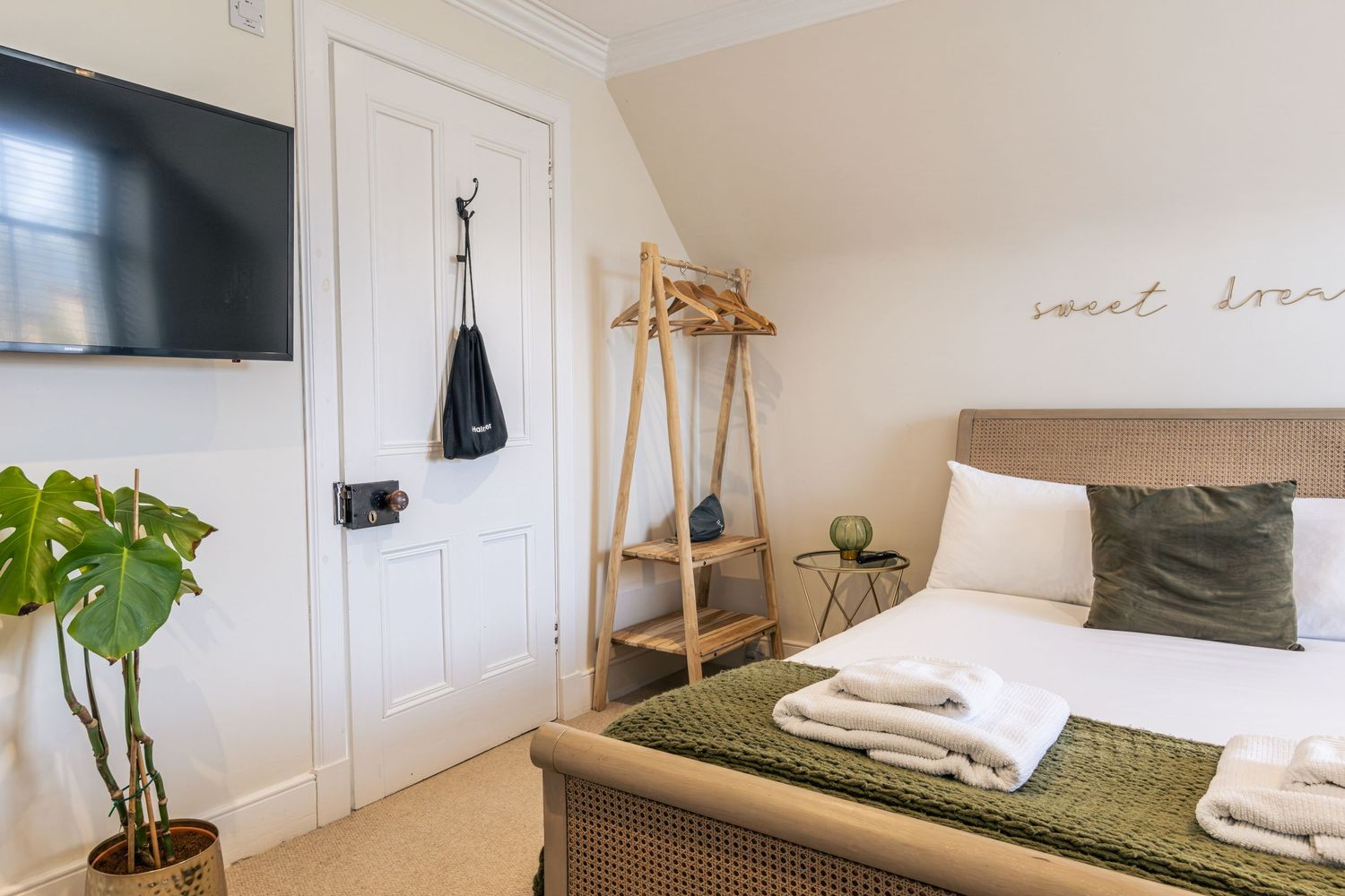 Cozy bedroom with a double bed, green accents, wooden ladder shelf, wall-mounted TV, and monstera plant.