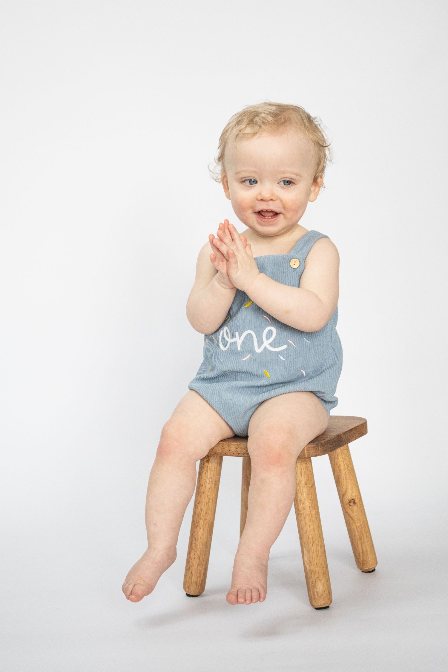 Smiling baby in blue 'one' romper sitting on wooden stool, clapping hands, white background, first birthday photo.