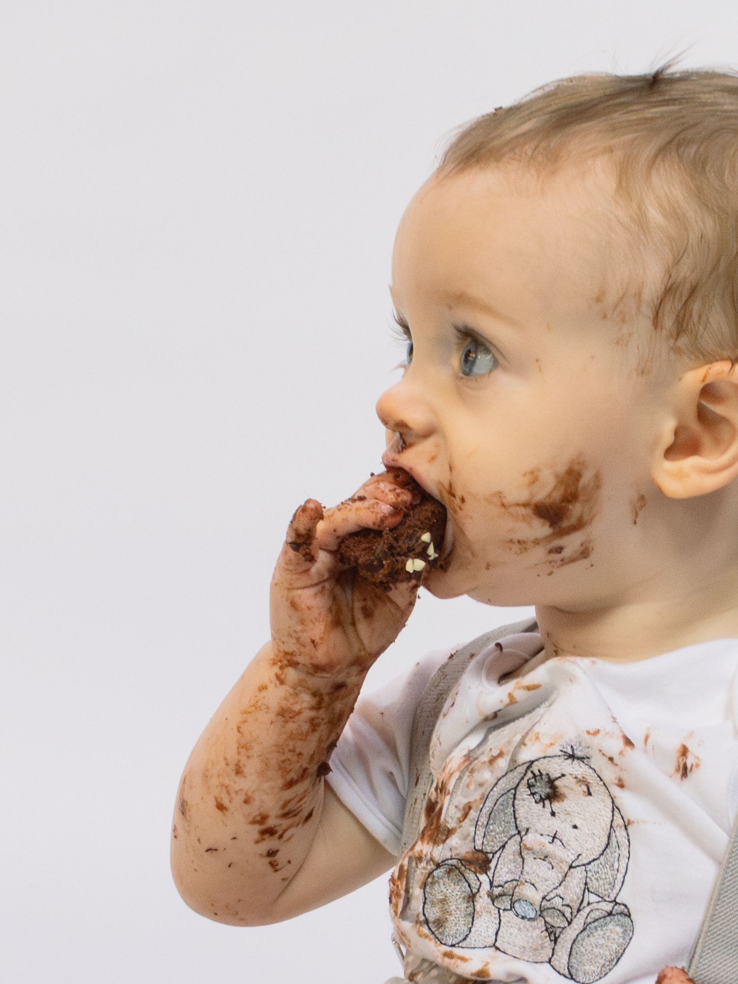 A messy toddler with blue eyes enjoys chocolate cake, with smears covering their face, hands, and white bear-print shirt.