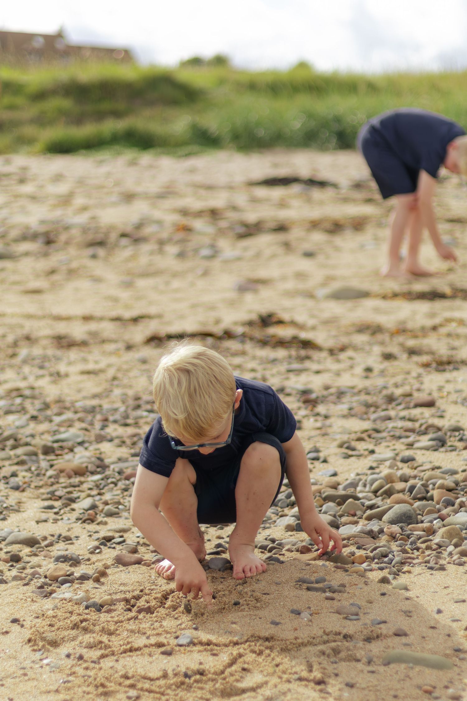 Young blonde boy crouching on a sandy beach collecting pebbles.