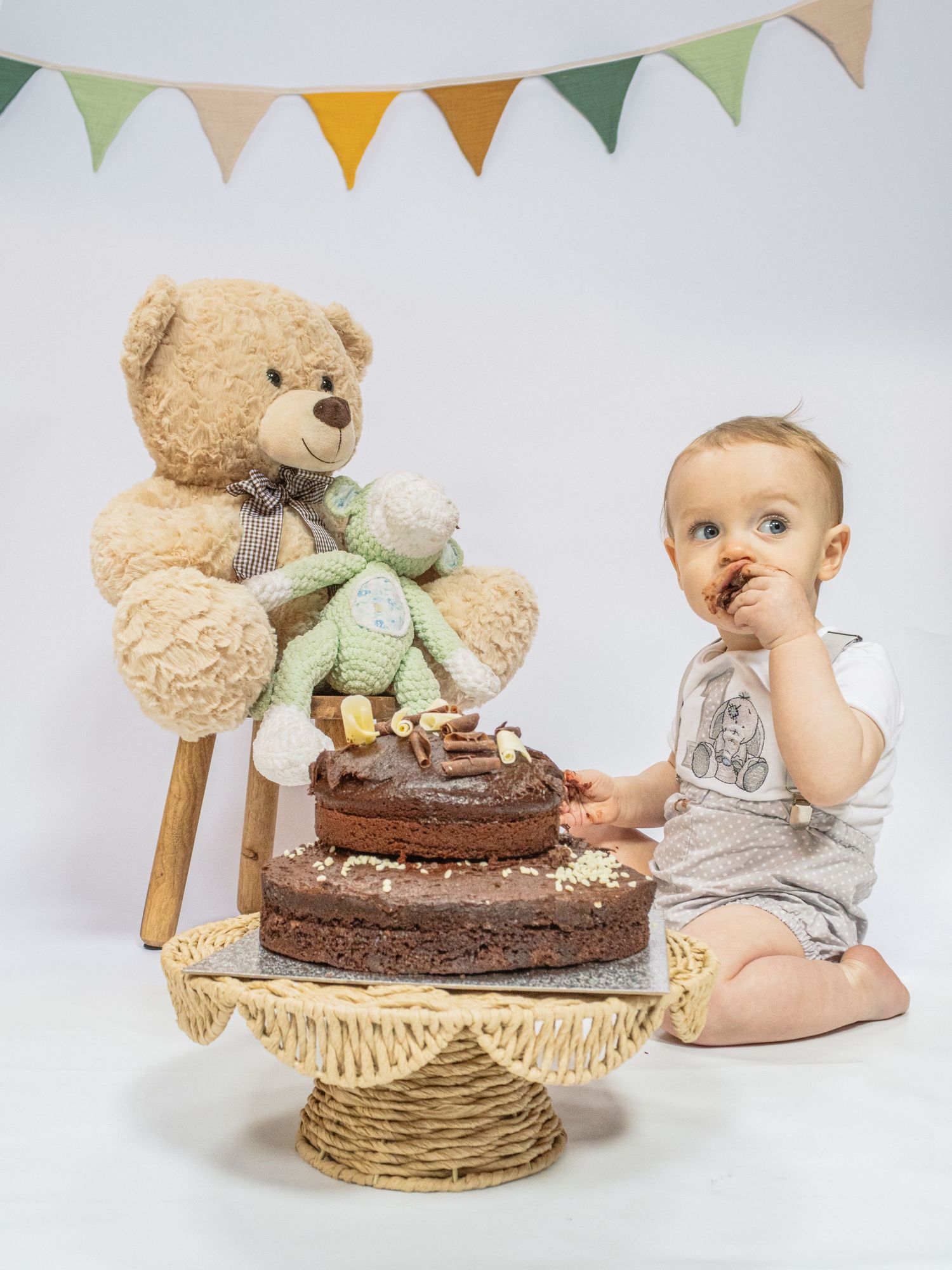 Baby boy eating chocolate smash cake beside teddy bear and green frog toy with bunting flags backdrop.