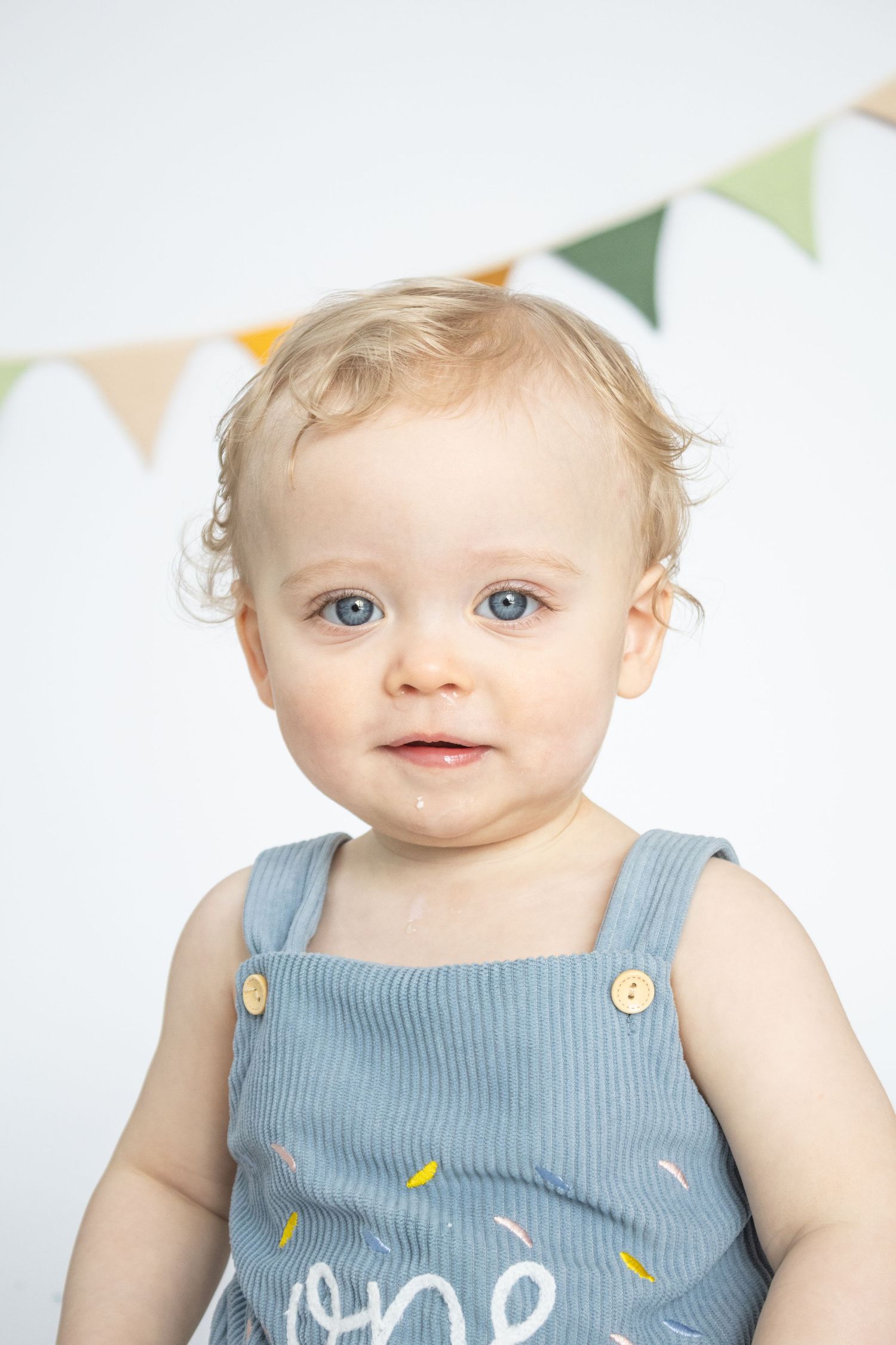 Adorable baby with blue eyes wearing a blue corduroy romper sits in front of colorful bunting flags.