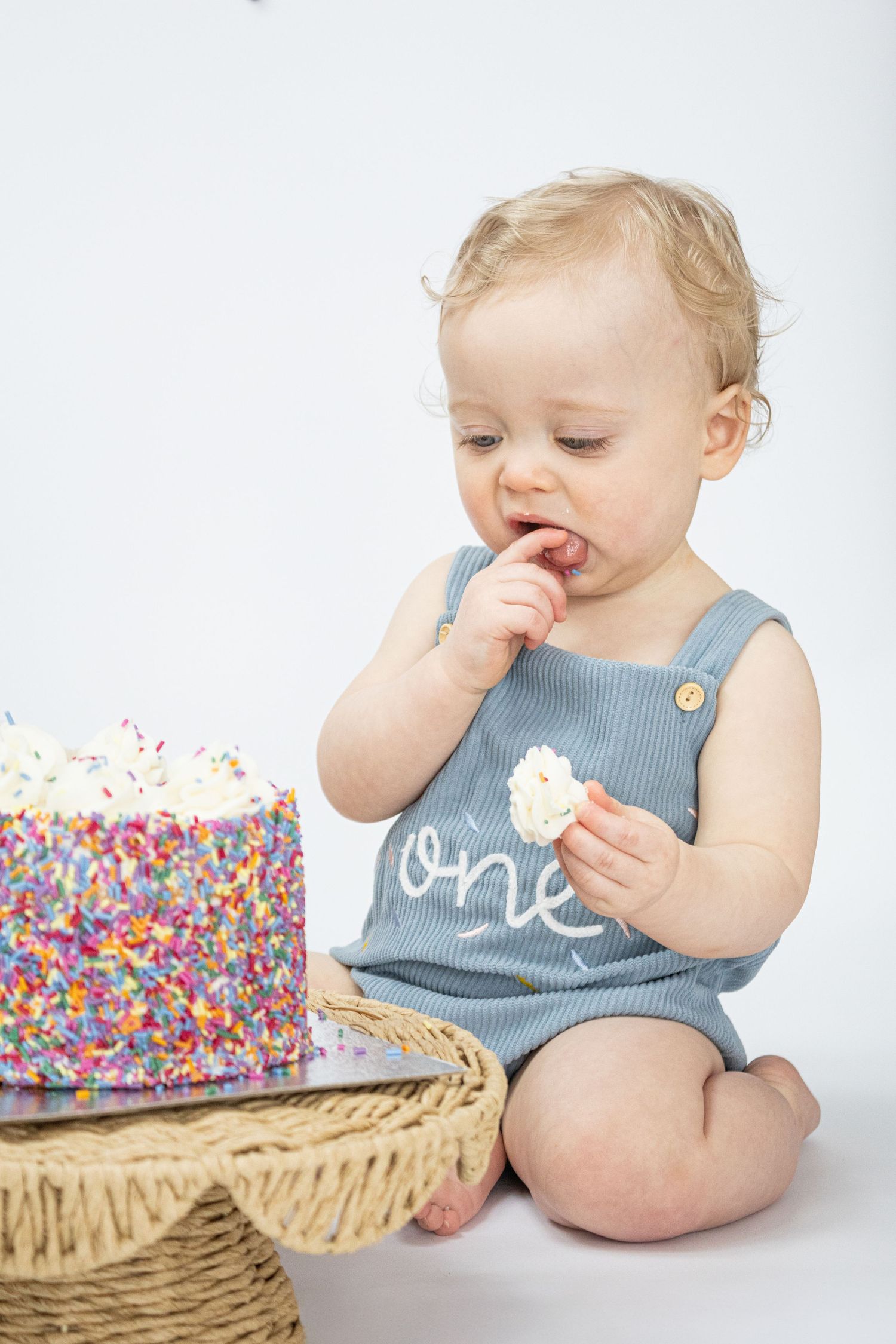 Baby in a blue “one” romper eating cake at a first birthday smash cake session.