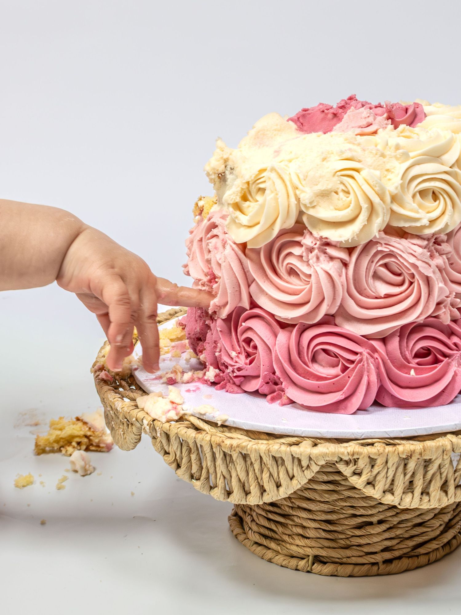 A baby hand reaching into a pink ombre rosette birthday cake on a wicker stand with crumbs scattered around.