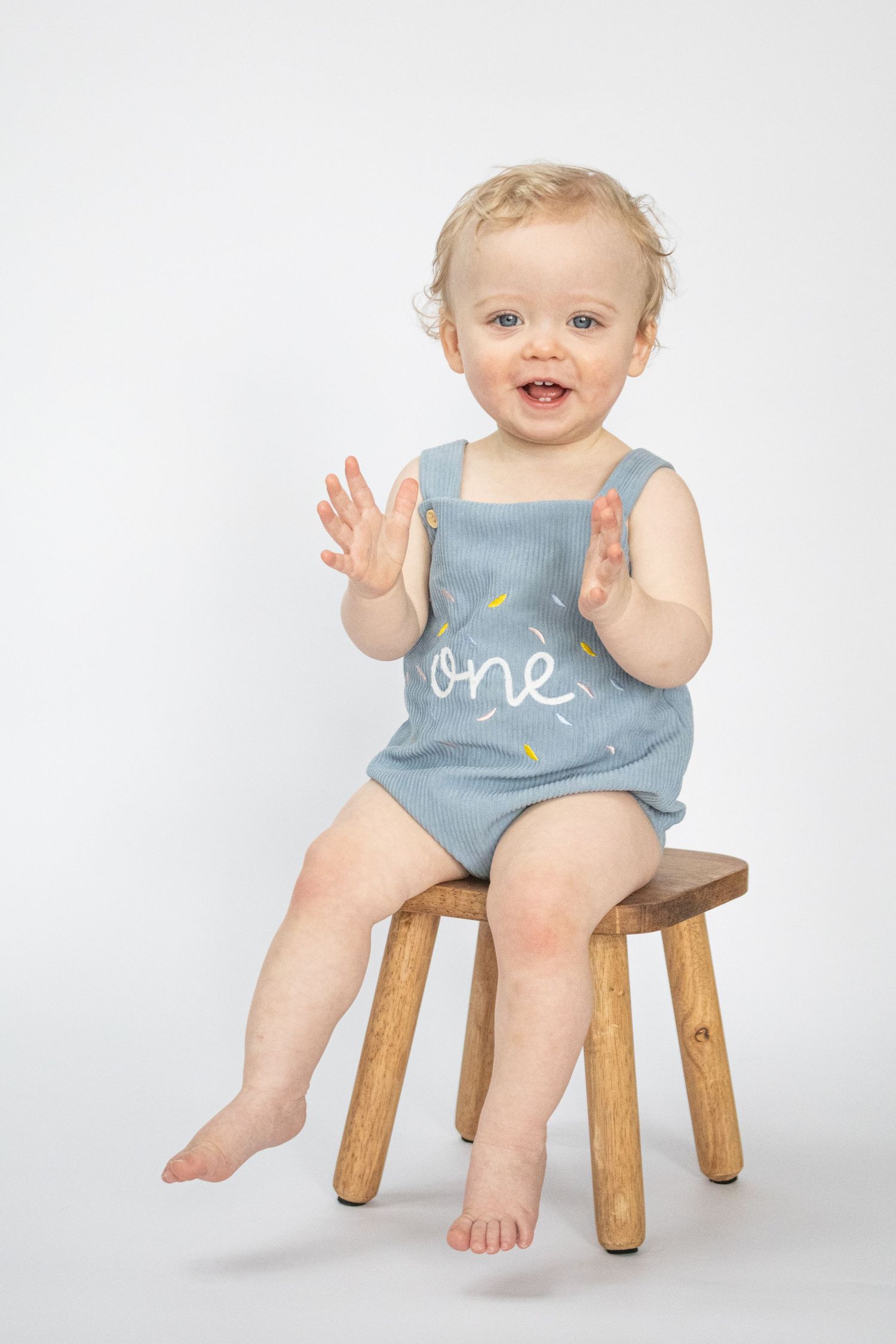 Smiling one-year-old baby clapping hands, wearing blue romper with 'one' text, sitting on wooden stool against white background.