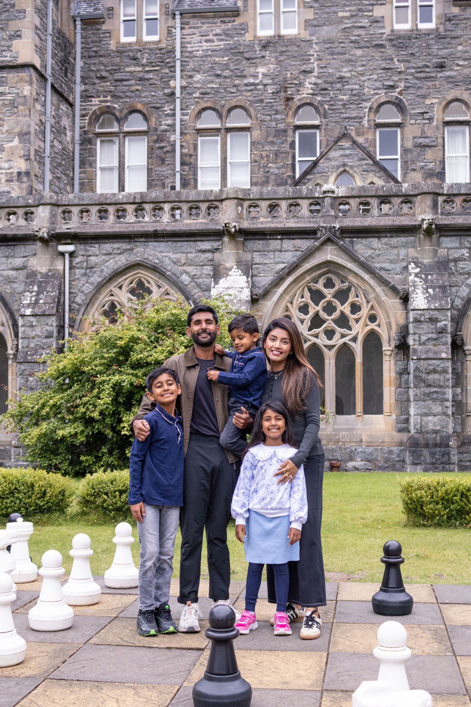 Family of five posing happily in front of a Gothic stone building with giant chess pieces on the ground.