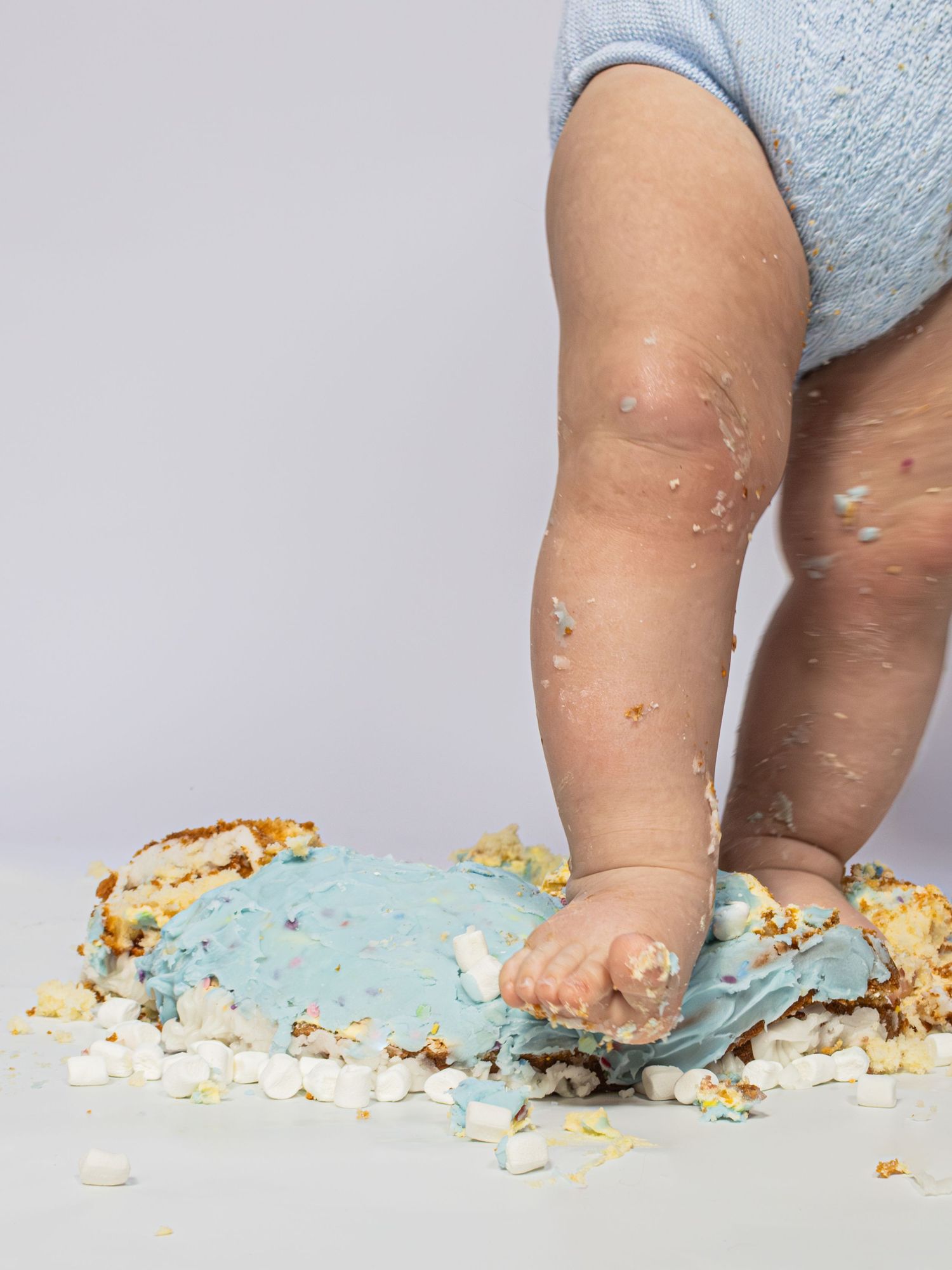 Baby feet stepping on smashed blue birthday cake during a cake smash photoshoot on white background.