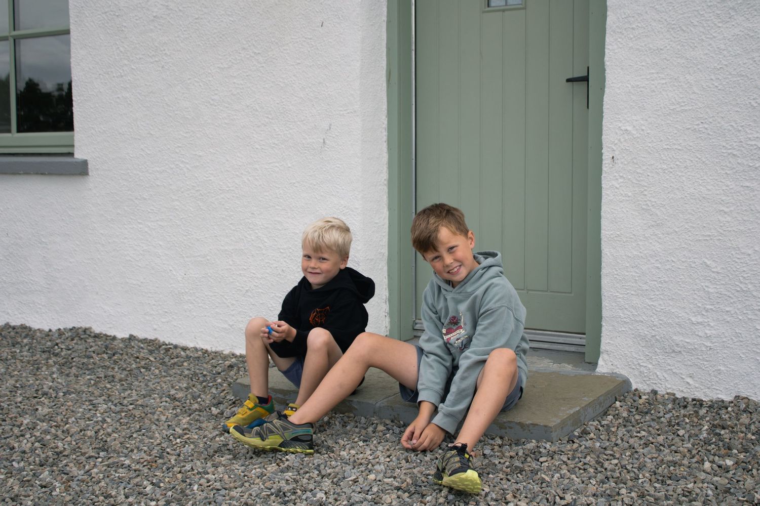 Two young children sit on a doorstep tying their shoes against a white wall with a mint green door.