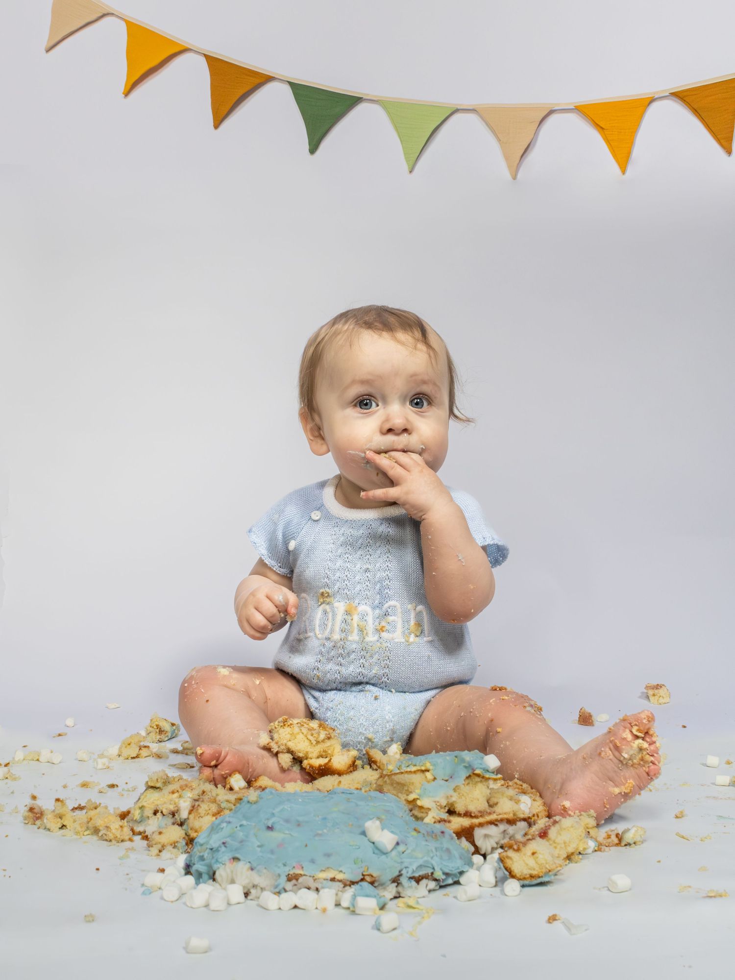 Baby boy in blue onesie smashing first birthday cake on floor with colorful bunting banner above.