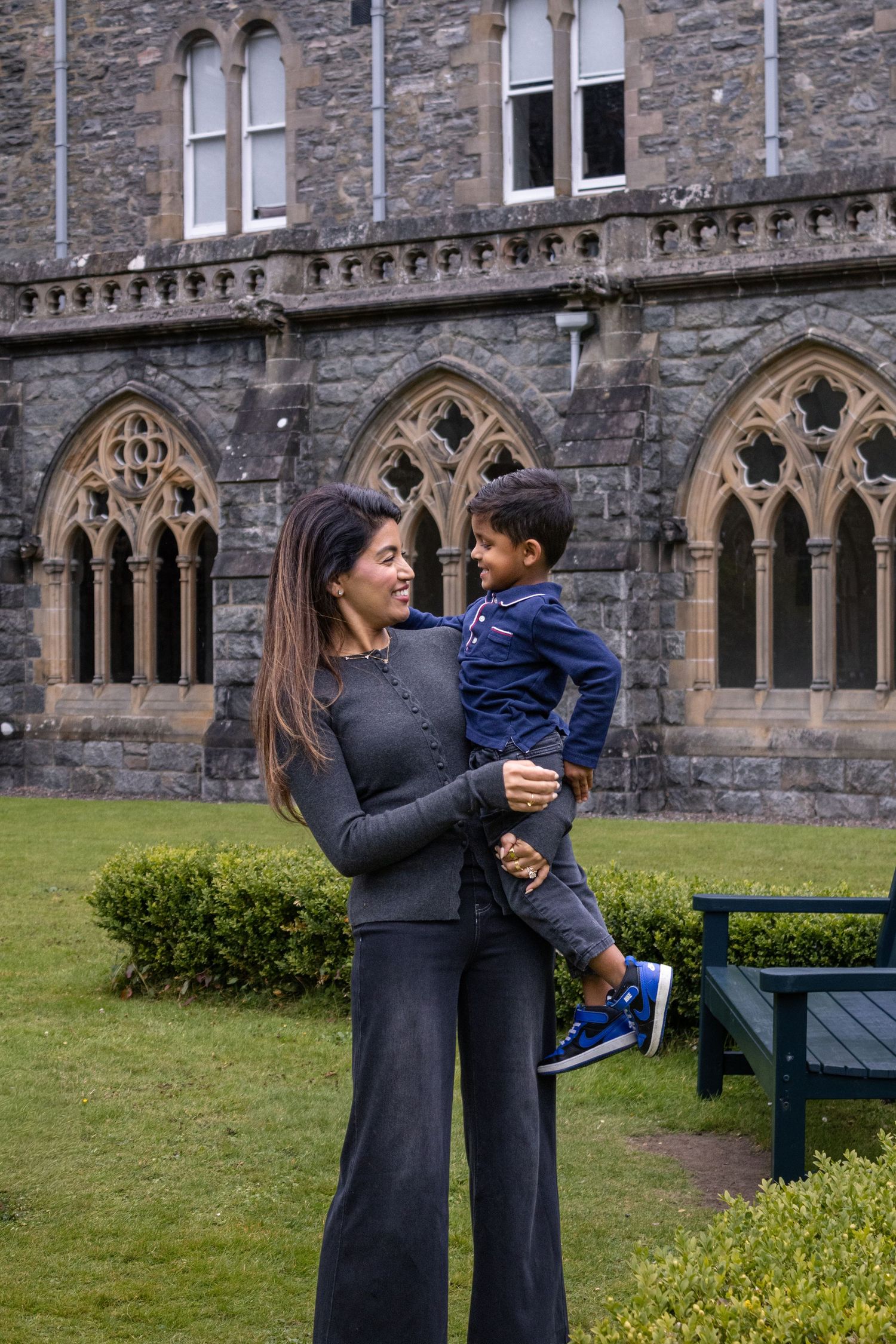 A woman holds a smiling toddler outside a historic stone Gothic building with arched windows and green lawn.