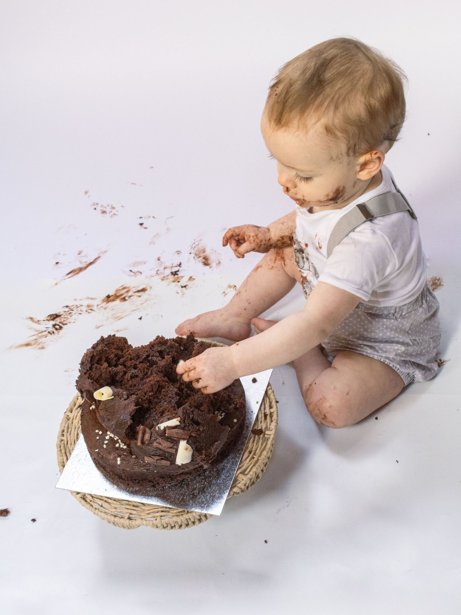 Toddler smashing chocolate birthday cake, covered in frosting, sitting on wicker basket mat.