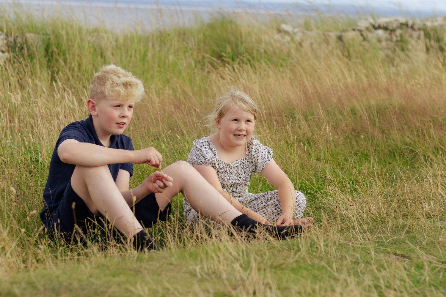 Two children sitting together in tall golden grass near a stone wall, enjoying a sunny outdoor day.