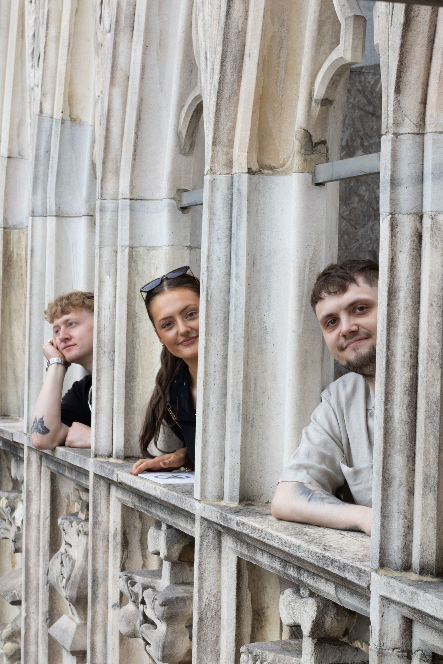 Three people peek playfully through stone archways of a historic building's architectural details.