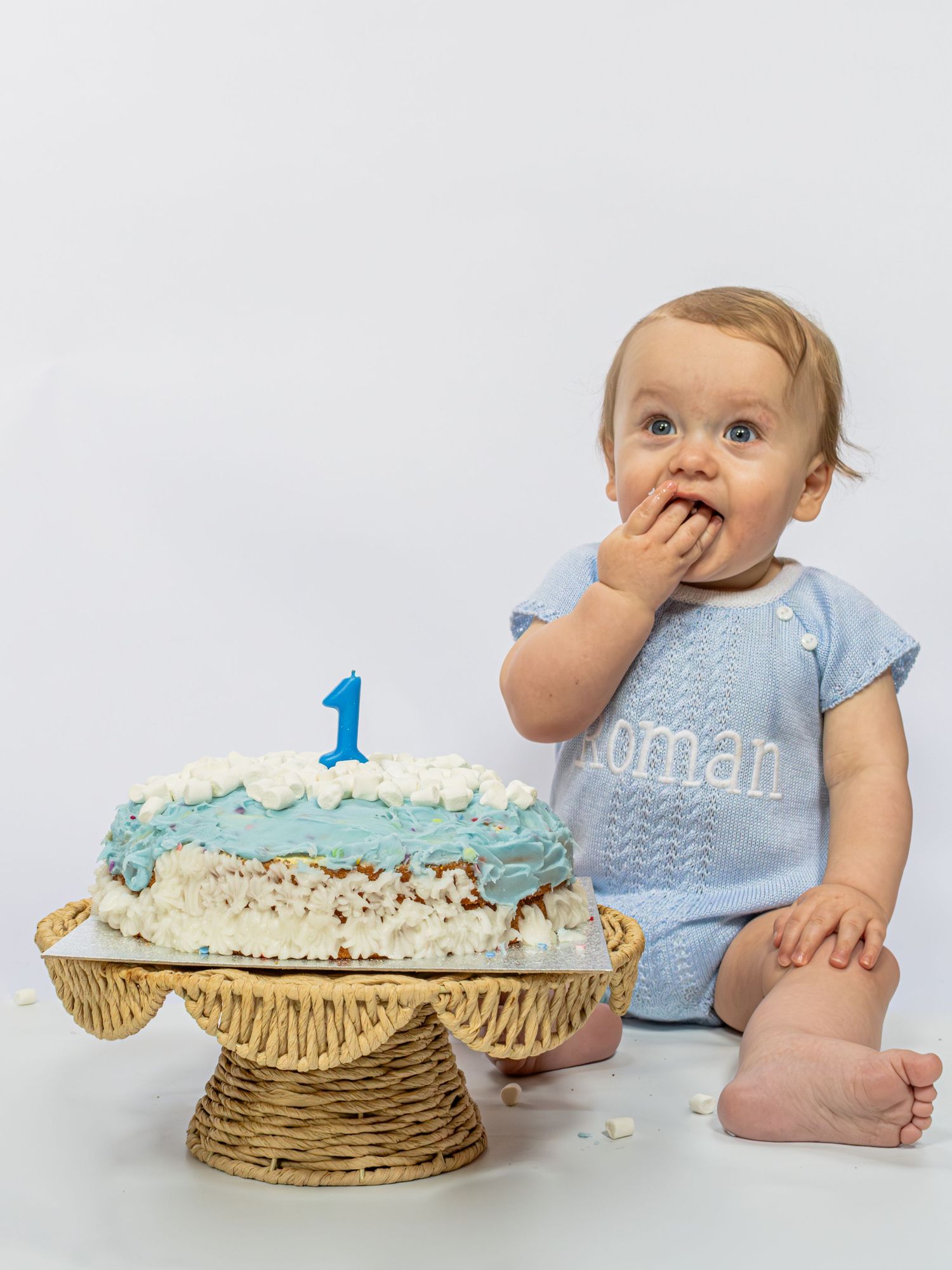 Baby boy in blue romper enjoys first birthday cake smash with blue number one candle on wicker stand.