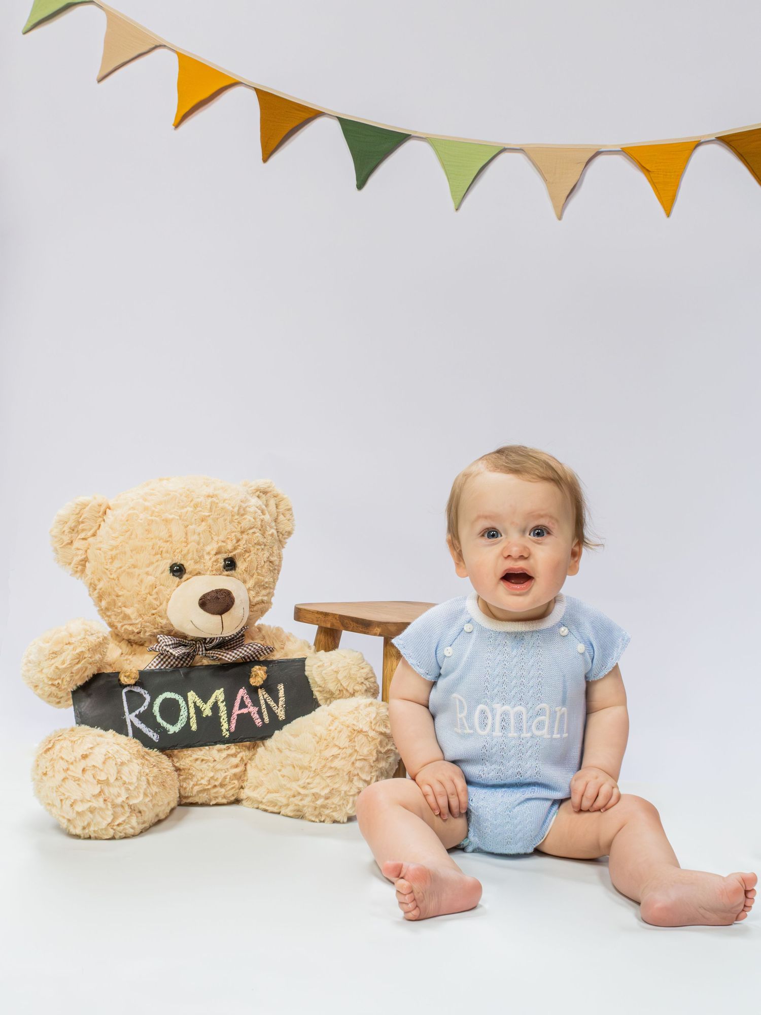 Baby Roman sits smiling beside a teddy bear holding a chalkboard sign, wearing a monogrammed blue romper under colorful bunting.
