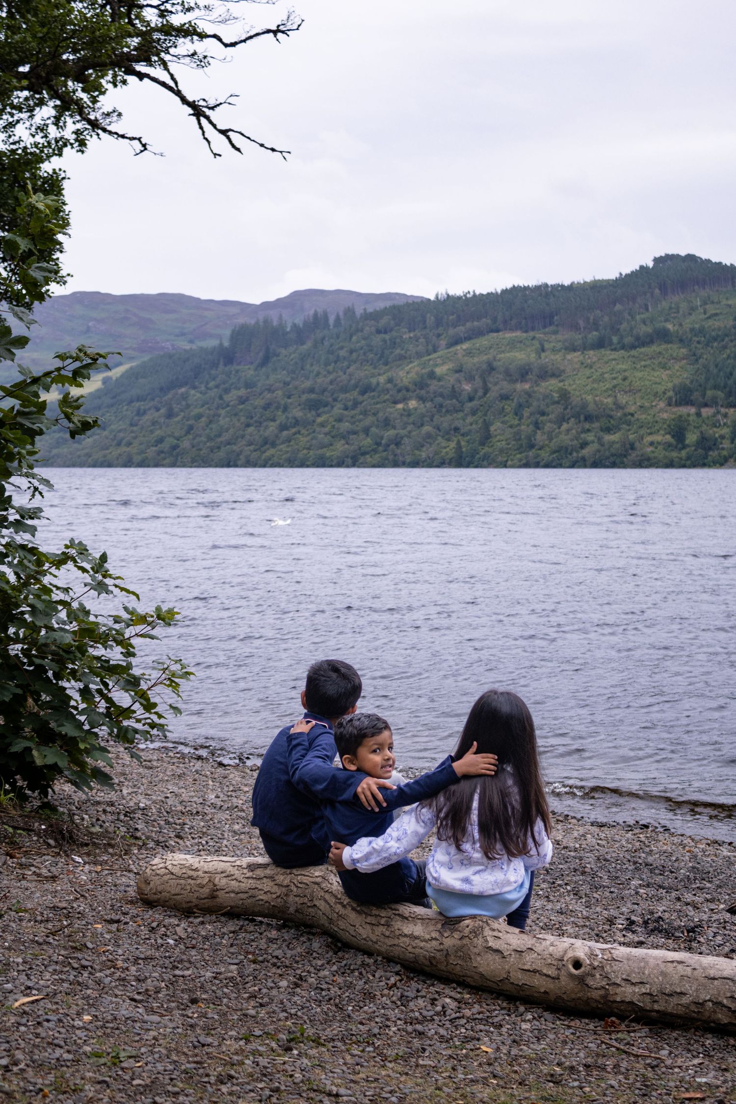 Three children sit on a log by a scenic loch, surrounded by green hills and trees on a cloudy day.