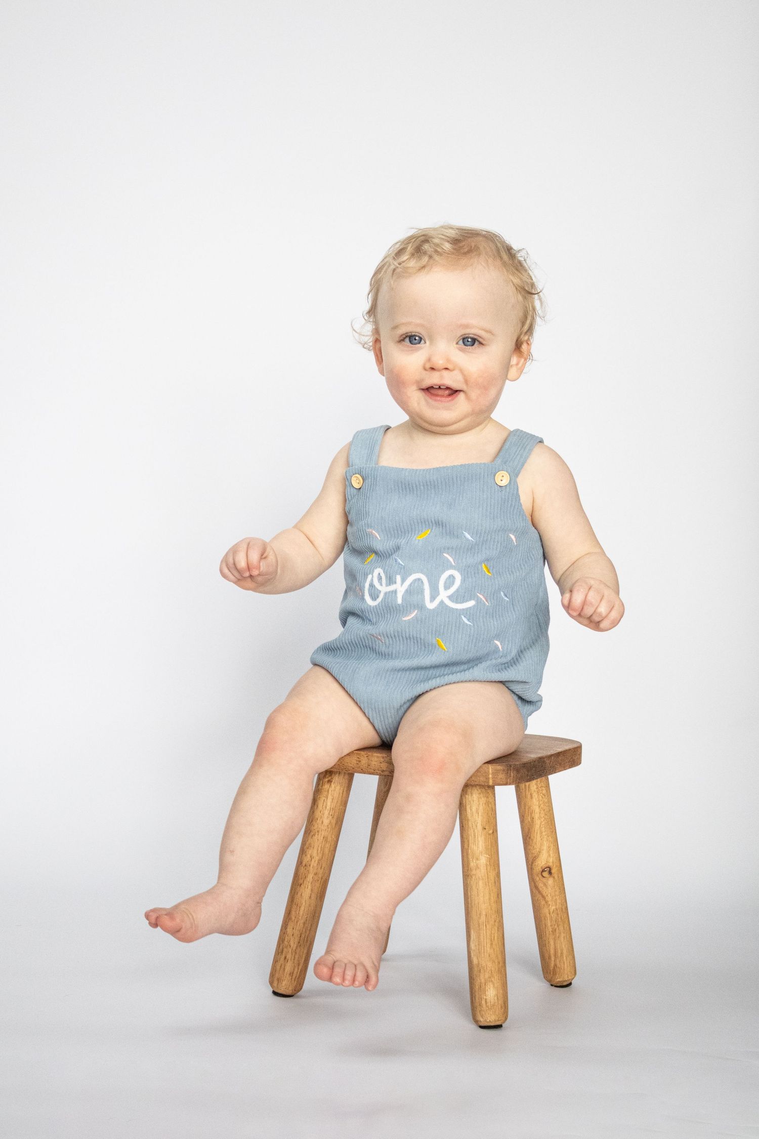 Smiling baby in blue 'one' romper sitting on wooden stool for first birthday portrait photo.