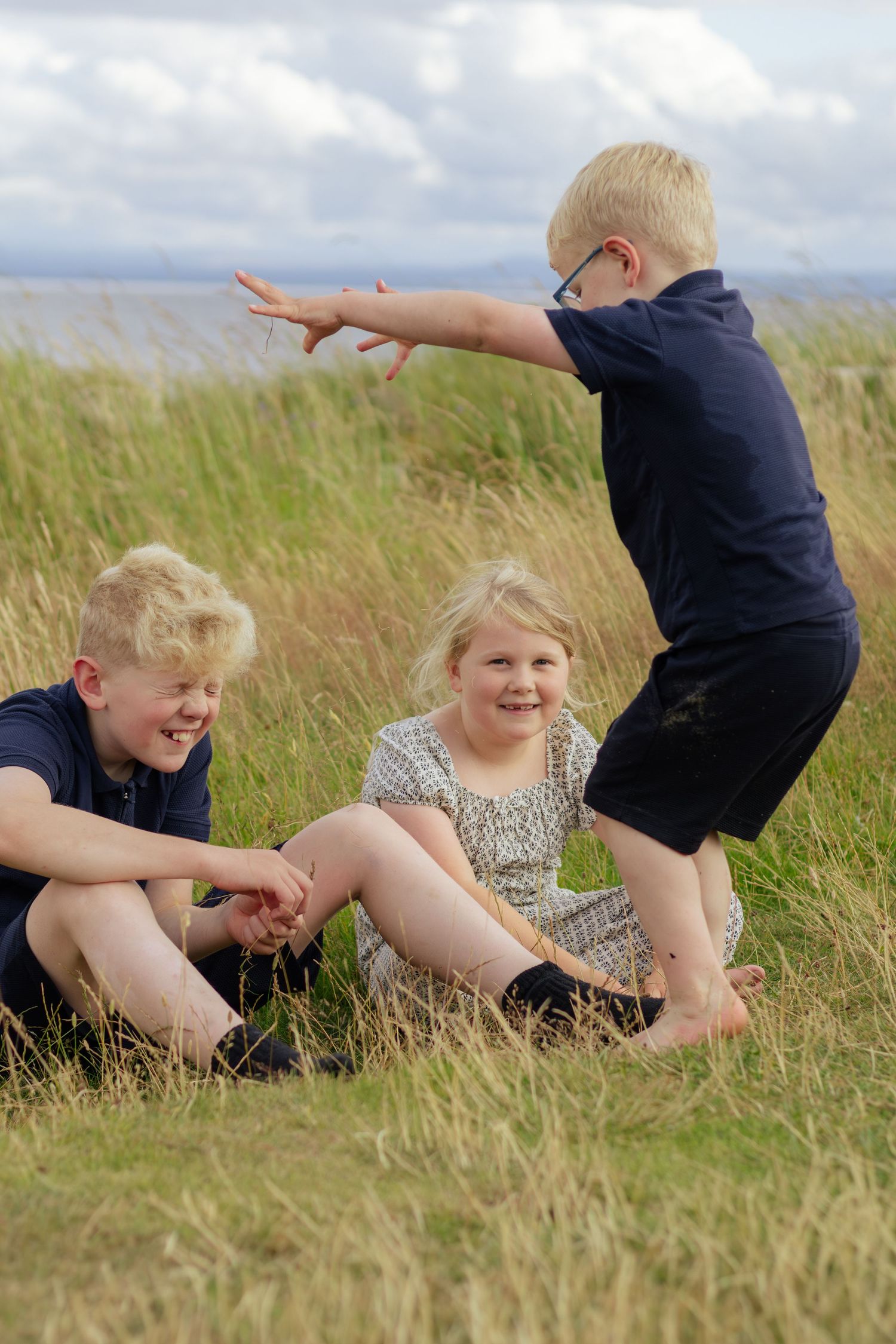 Three blonde children playing together in coastal grass on a cloudy day.