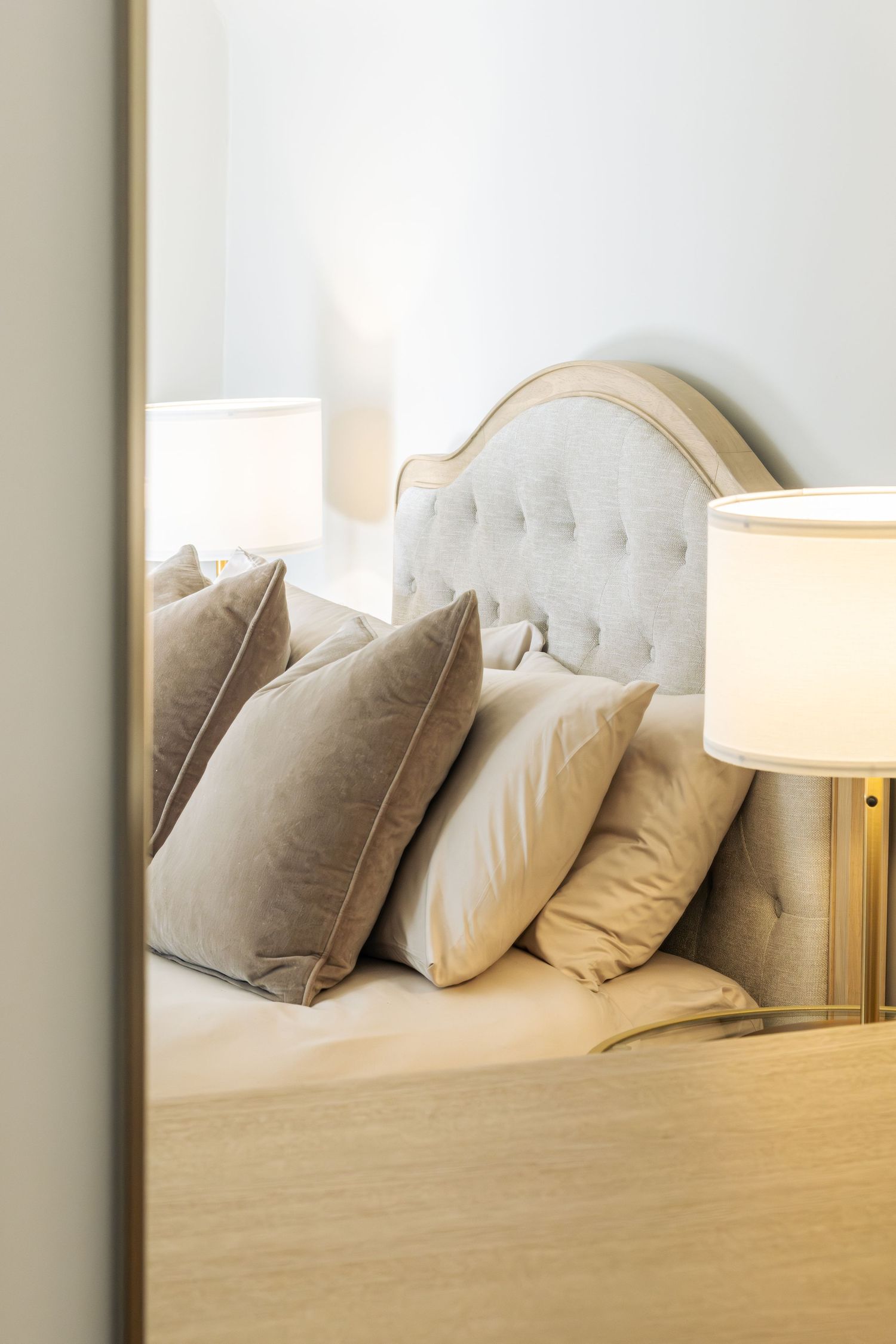 Elegant bedroom with tufted headboard, beige and taupe pillows, and warm glowing bedside lamp on wooden nightstand.