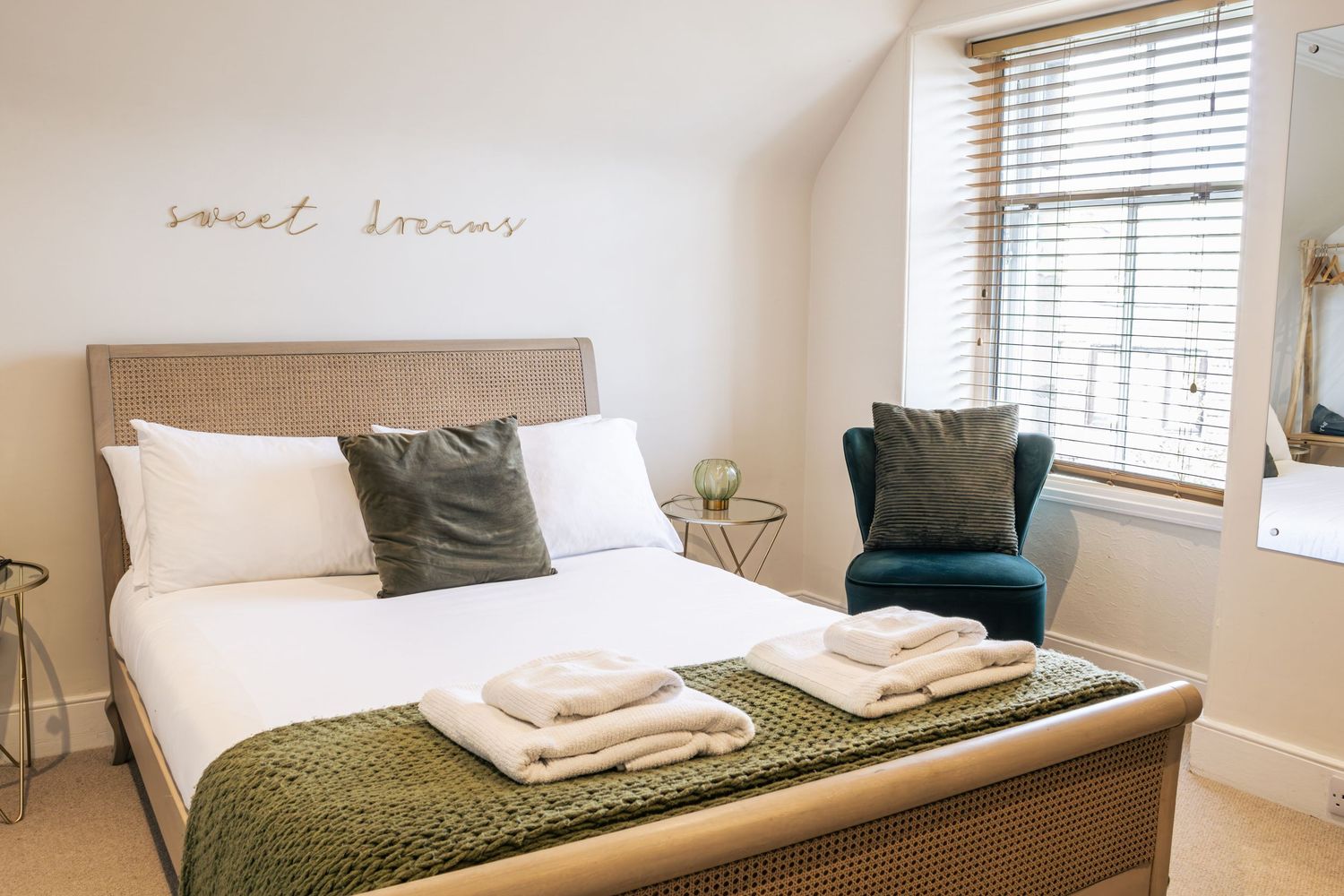 Elegant bedroom with a wooden bed frame, white bedding, green knit throw, teal armchair, and sweet dreams wall art.