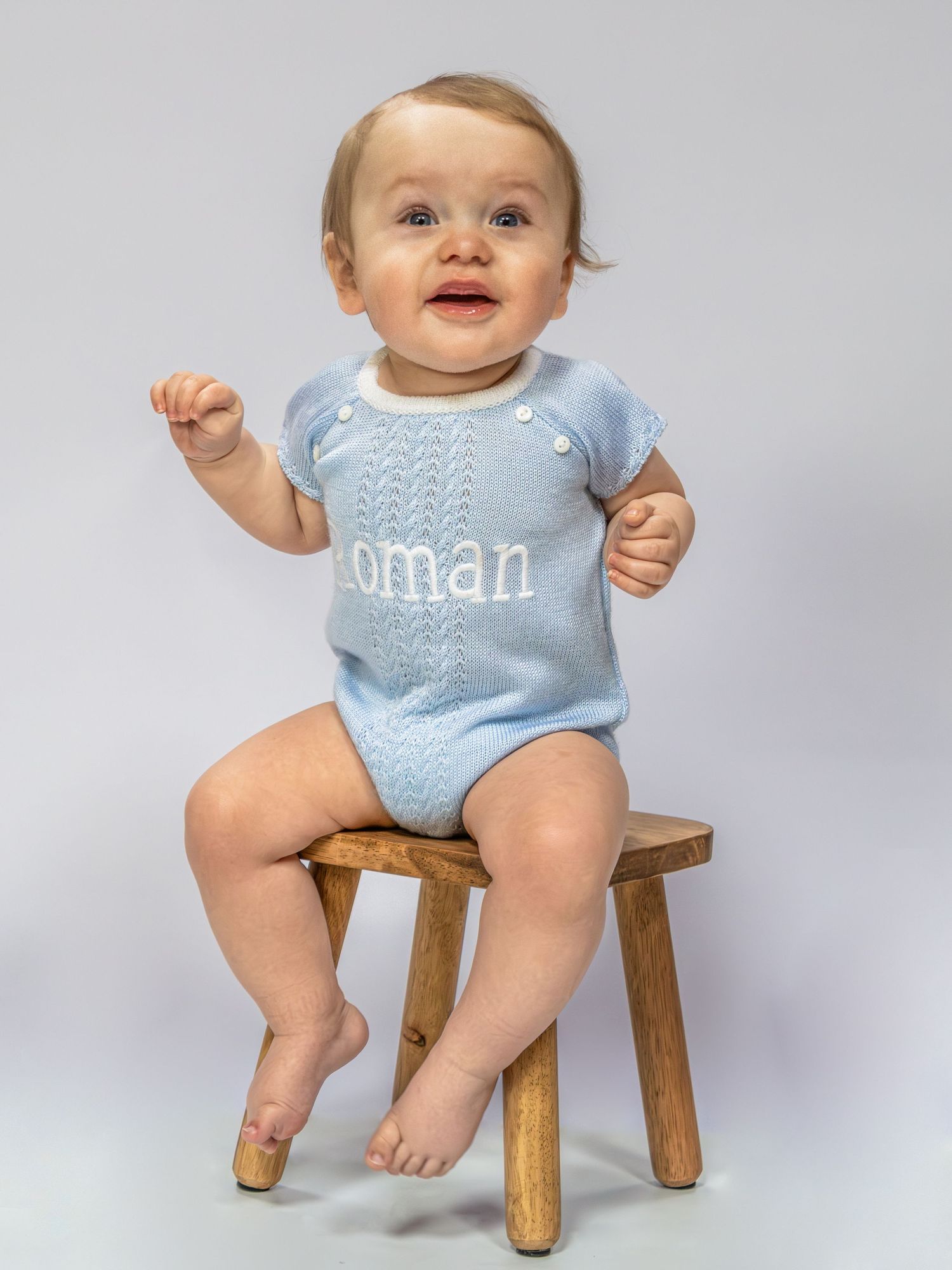 Baby boy smiling in a light blue knit personalised romper with name, seated on a wooden stool.