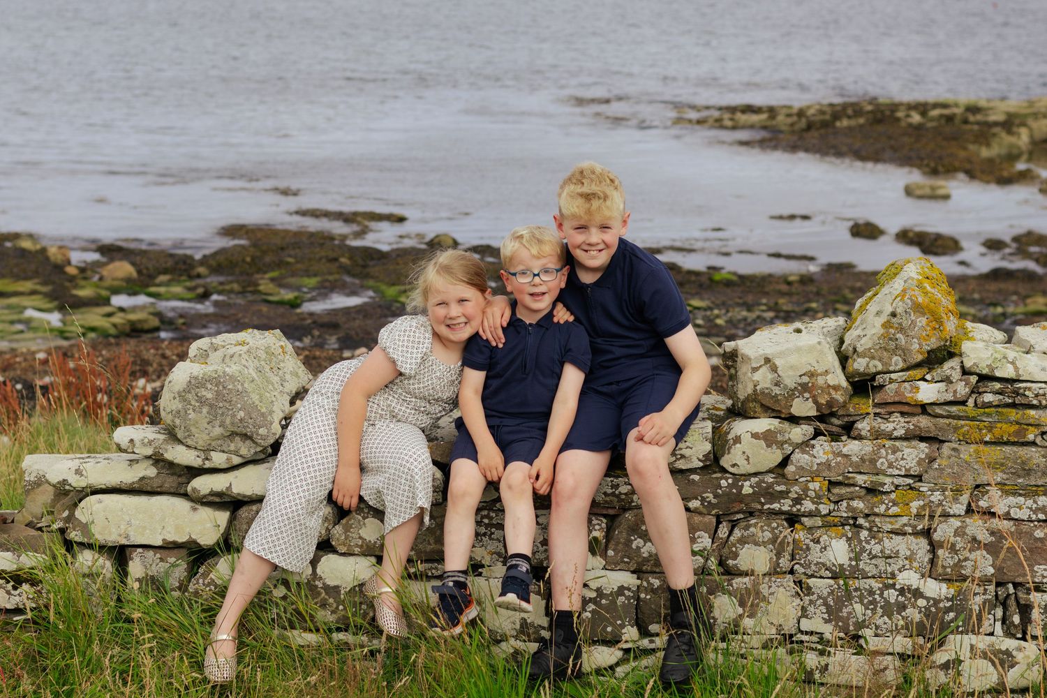 Three smiling blonde children sitting together on a stone wall by a scenic coastal shoreline.