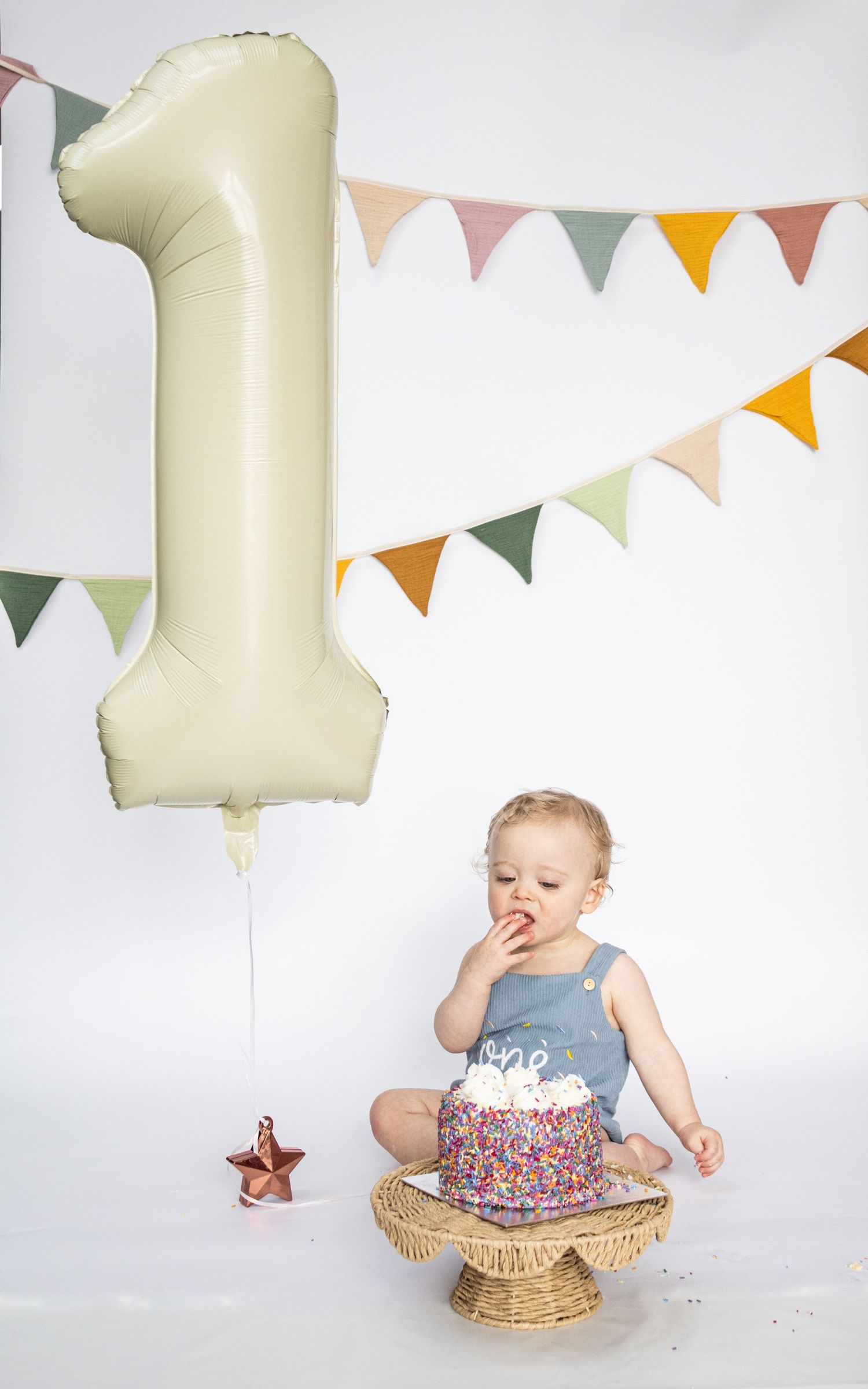 Baby celebrating first birthday with sprinkle cake, number one balloon, and colorful bunting banner.