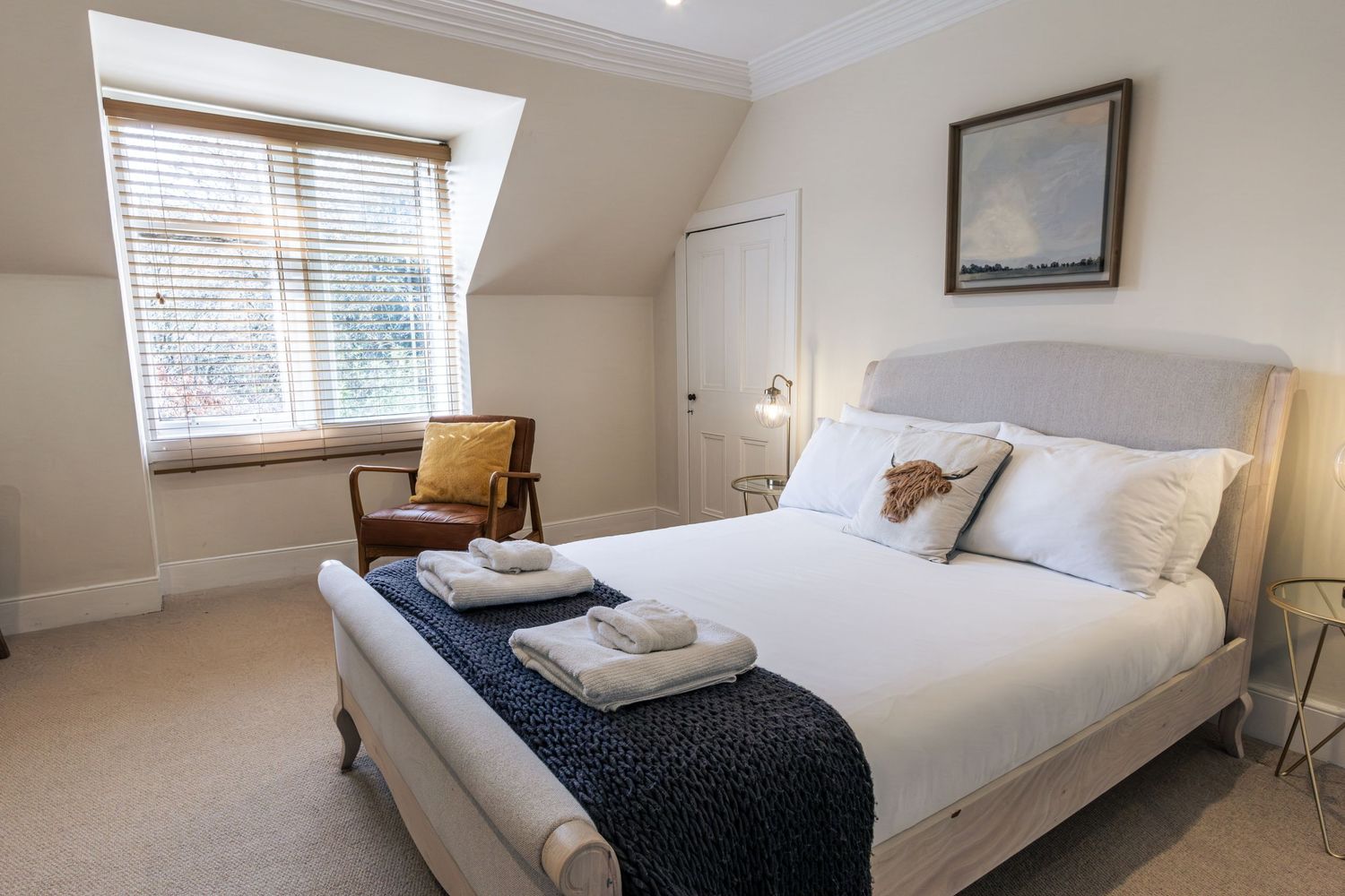Elegant white bedroom with a large bed, navy throw, skylight window, and a cozy armchair in a bright, modern setting.