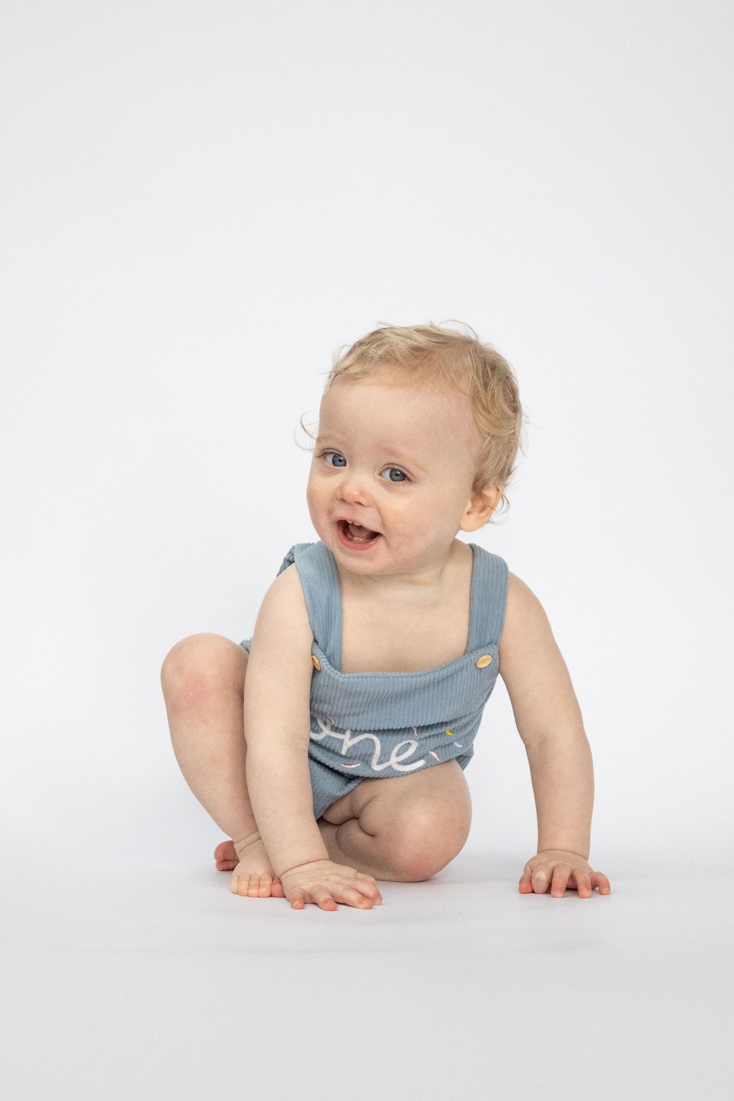 Happy baby in blue romper with 'one' text sits smiling on white background, celebrating first birthday.