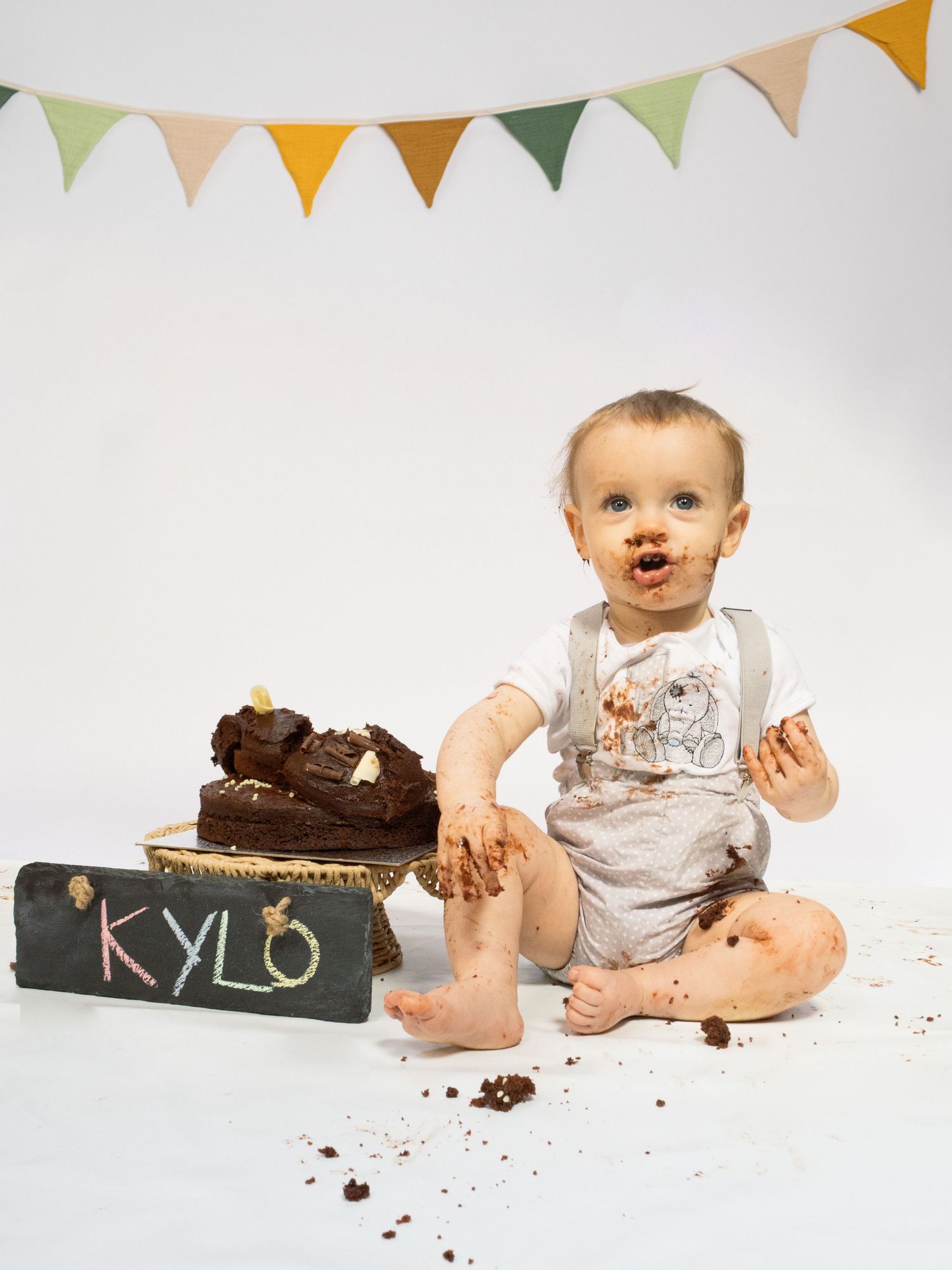 Baby Kylo covered in chocolate cake during first birthday smash cake photoshoot with colorful bunting.