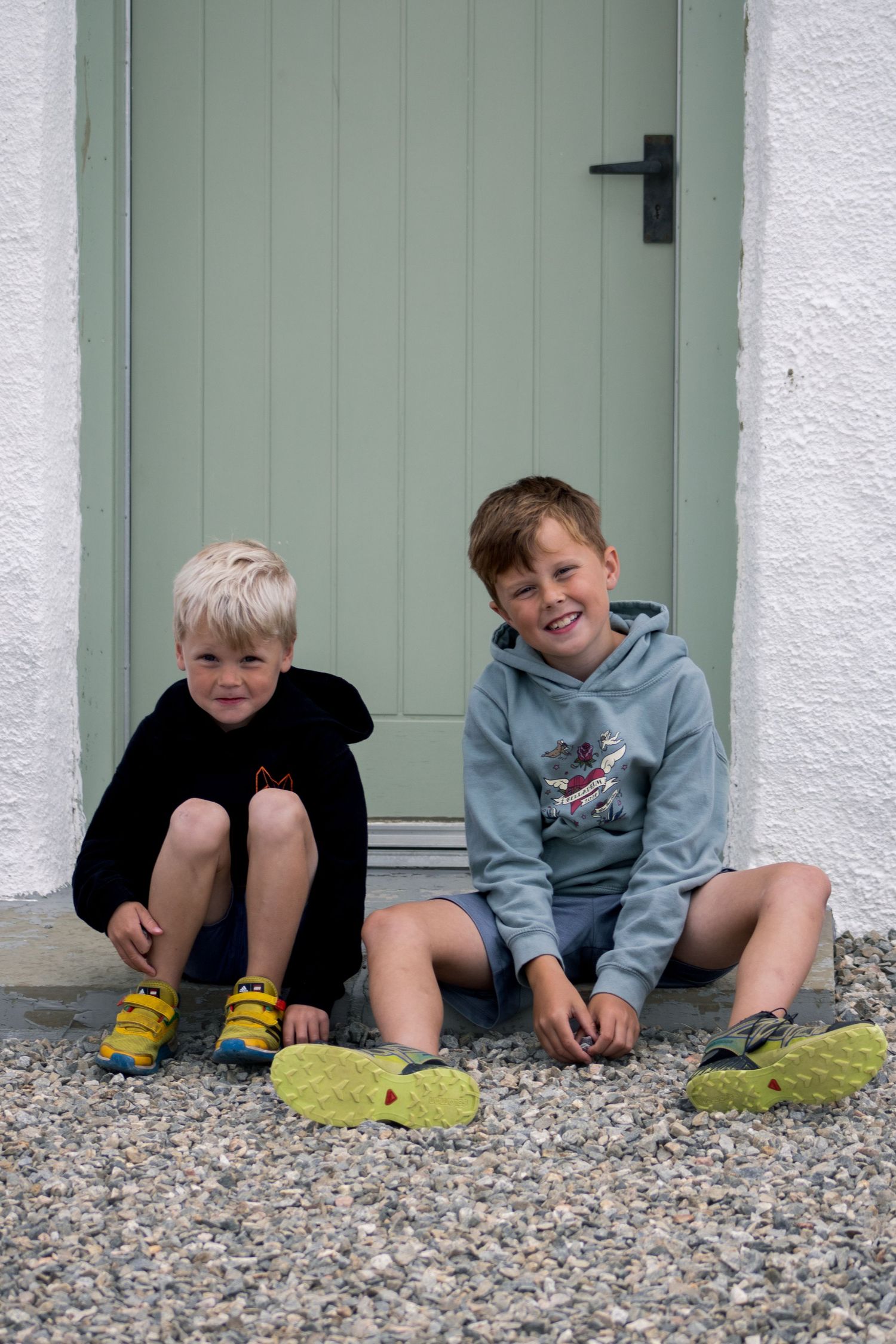 Young children lying down and laughing together on gravel ground next to a white wall with green doors.