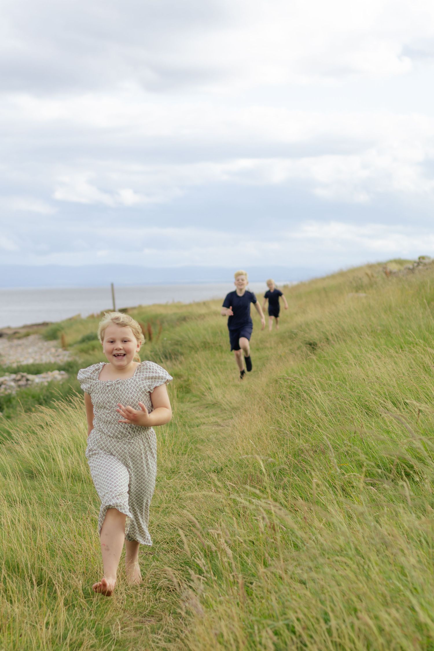 Children running through green coastal grass with the sea visible in the background on a cloudy day.