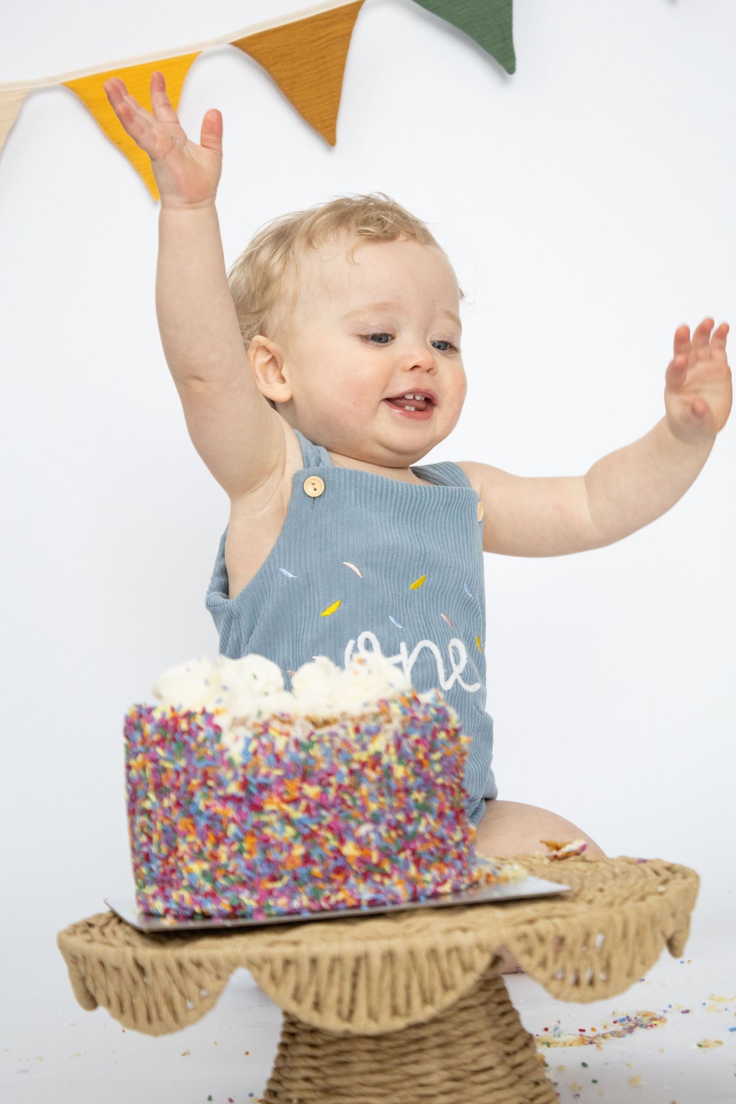 Excited baby boy raises hands over colorful sprinkle birthday cake during first birthday cake smash session.