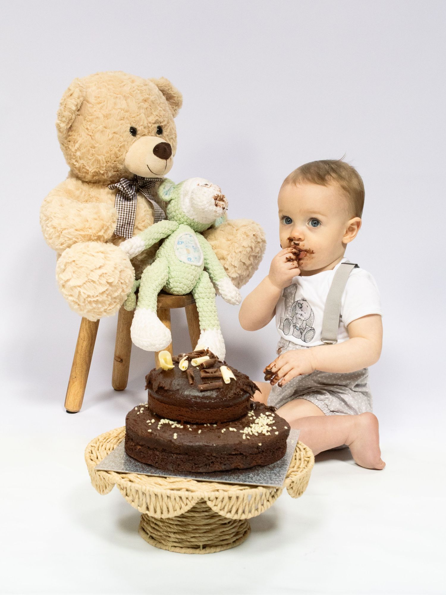 Toddler enjoying first birthday chocolate smash cake seated next to large teddy bear and frog stuffed toy.