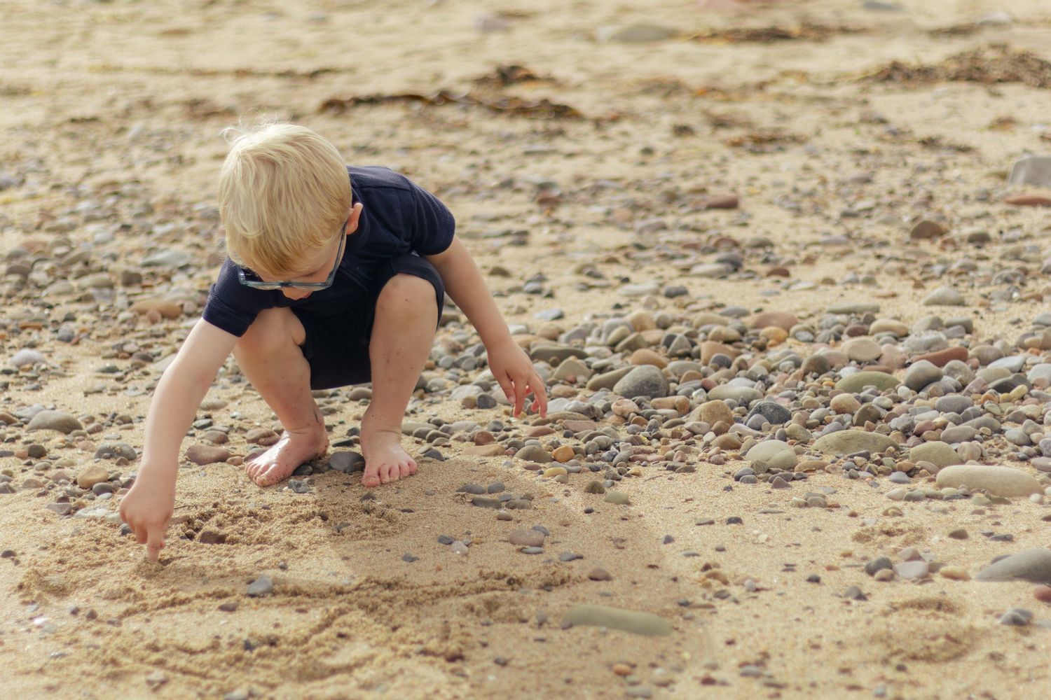 A young blonde toddler crouches on a sandy beach, playing with small pebbles and stones on a sunny day.