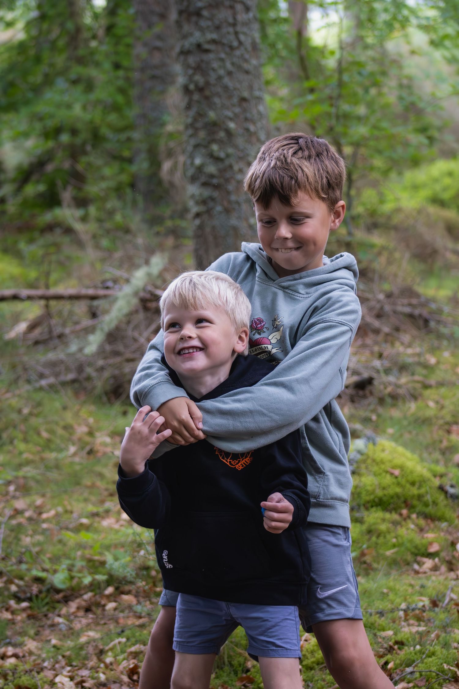 Two children playing and laughing together in a forest setting with green foliage.