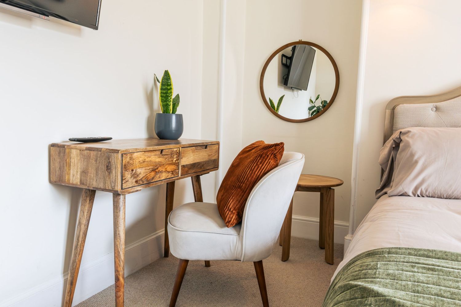Cozy bedroom corner with rustic wooden desk, white accent chair, round mirror, and potted succulent in a bright room.