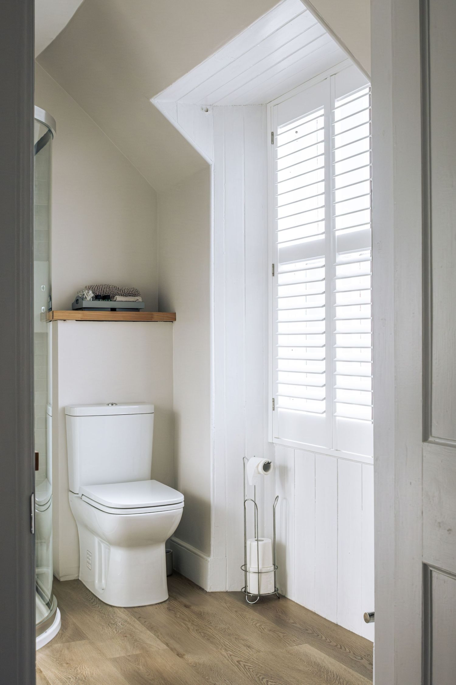 Bright white bathroom with toilet, wooden shelf, plantation shutters, and hardwood floors in a sloped ceiling alcove.