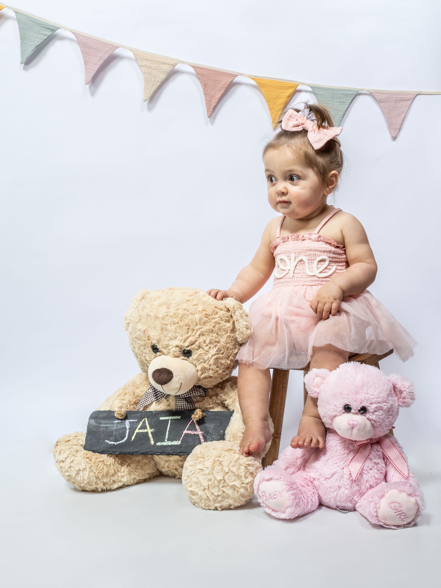 Baby girl in pink tutu dress poses with teddy bears and a name sign reading JAIA at a first birthday photo shoot.