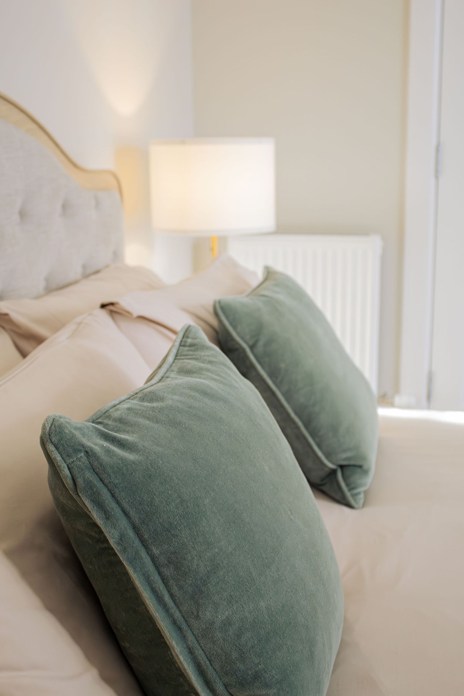 Elegant bedroom with two sage green velvet pillows on a white bed, featuring a gold lamp and tufted headboard.