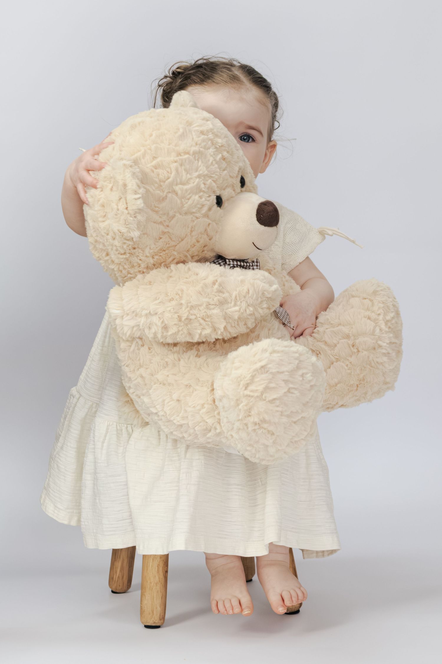 A little girl in a white dress hugging a large cream teddy bear while standing barefoot on a small wooden stool.