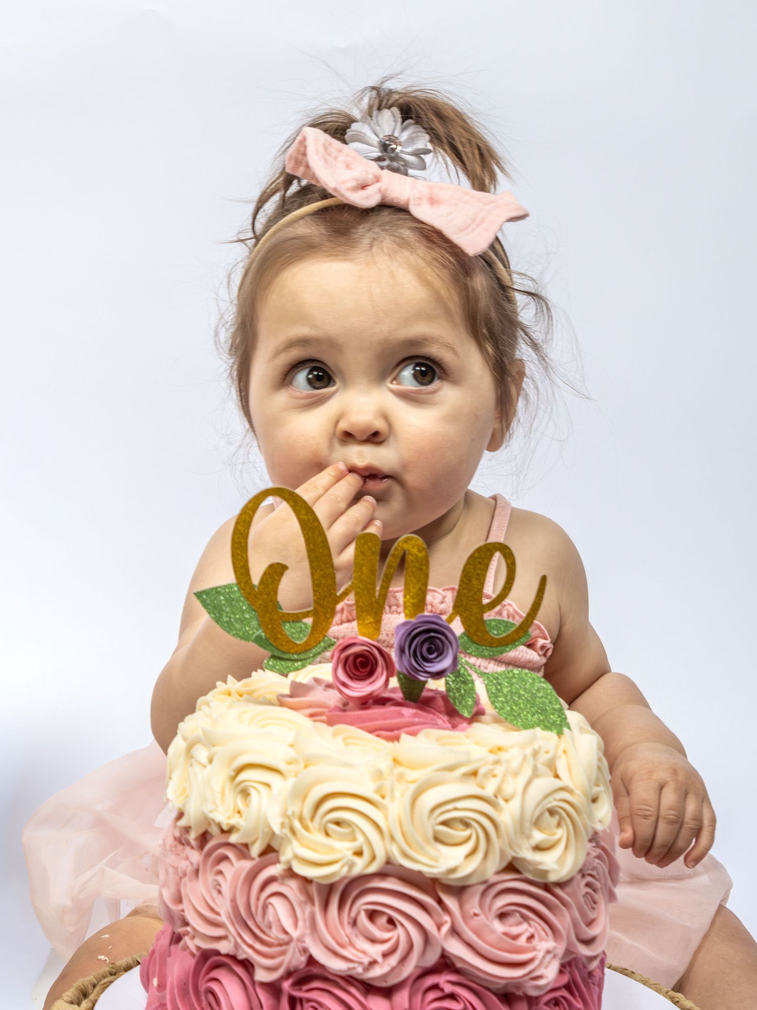 Adorable baby girl celebrating her first birthday with a pink rosette smash cake topped with a gold glitter One topper.